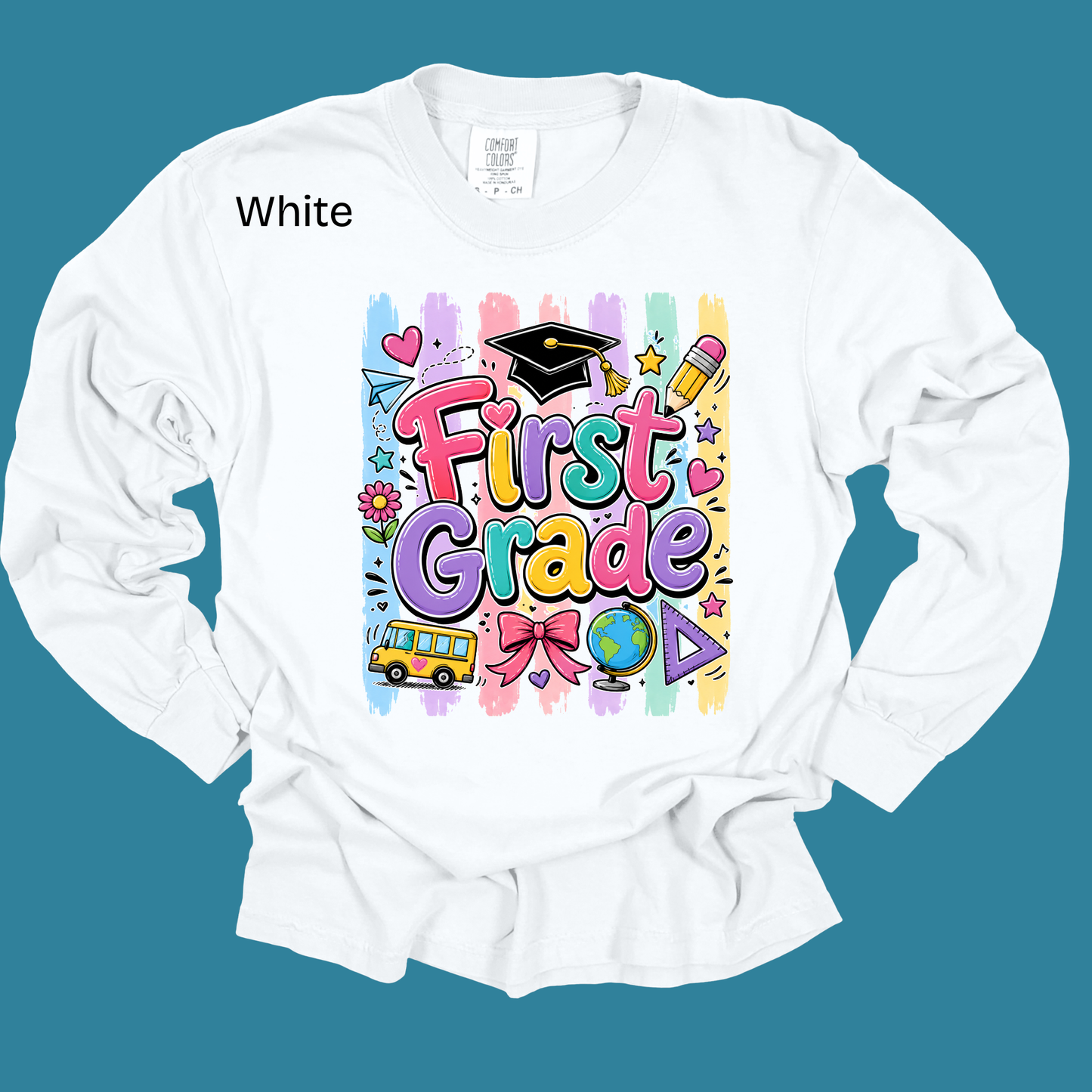 A white long-sleeve t-shirt with a colorful 'First Grade' graphic on the front, displayed against a solid teal background. The graphic features playful school-themed illustrations like a graduation cap, pencil, globe, and bus.