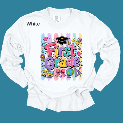 A white long-sleeve t-shirt with a colorful 'First Grade' graphic on the front, displayed against a solid teal background. The graphic features playful school-themed illustrations like a graduation cap, pencil, globe, and bus.