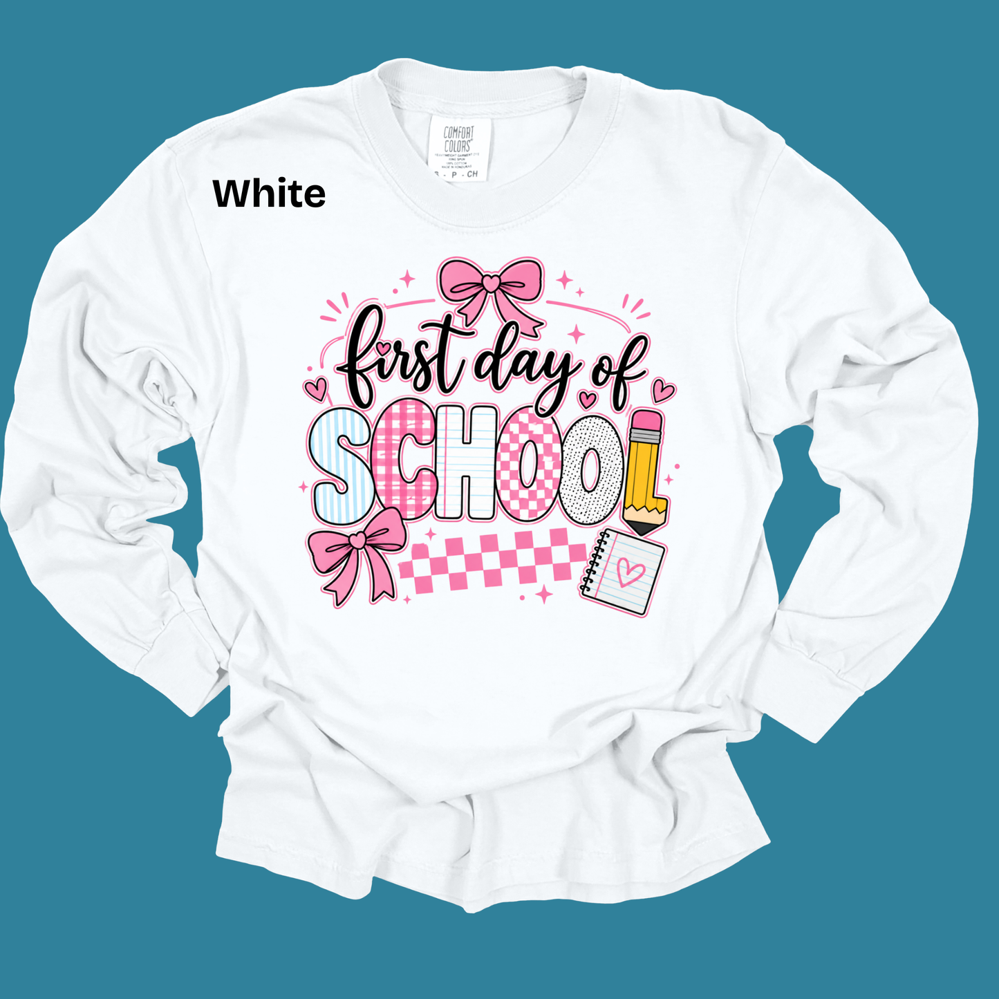 A white long-sleeve t-shirt with a 'first day of SCHOOL' graphic on the front, featuring pink bows, checkered patterns, and school-themed illustrations, shown against a solid teal background with 'White' text in the upper left.