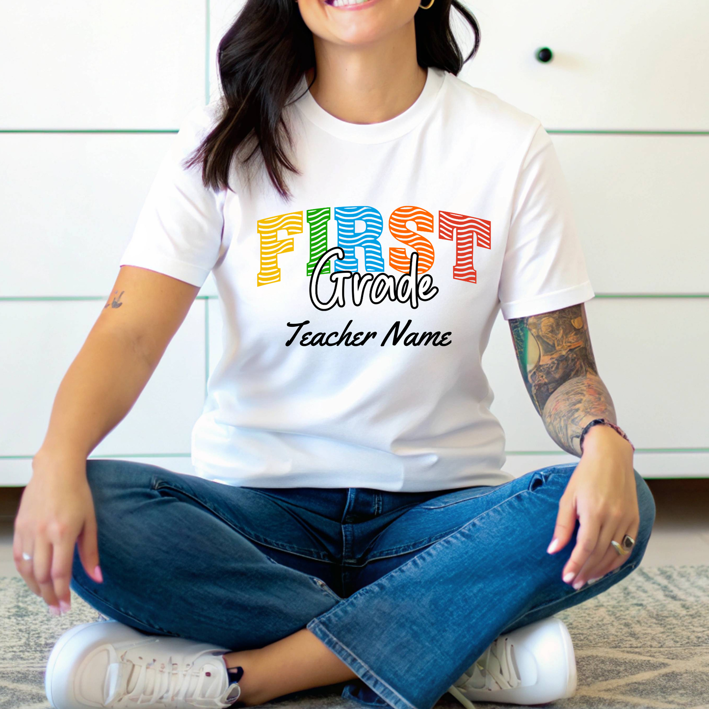 A woman sitting cross-legged wearing a white graphic t-shirt with 'FIRST Grade Teacher Name' printed in colorful retro letters, paired with blue jeans and white sneakers.