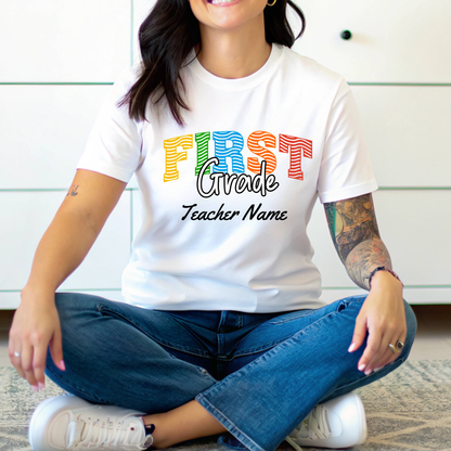 A woman sitting cross-legged wearing a white graphic t-shirt with 'FIRST Grade Teacher Name' printed in colorful retro letters, paired with blue jeans and white sneakers.