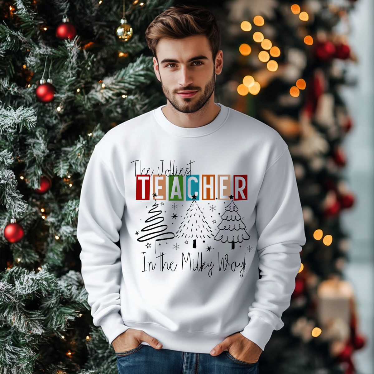A man wearing a white crewneck sweatshirt with a colorful graphic that reads 'The Jollyest TEACHER In The Miley Way' with Christmas tree illustrations, standing in front of a decorated Christmas tree.