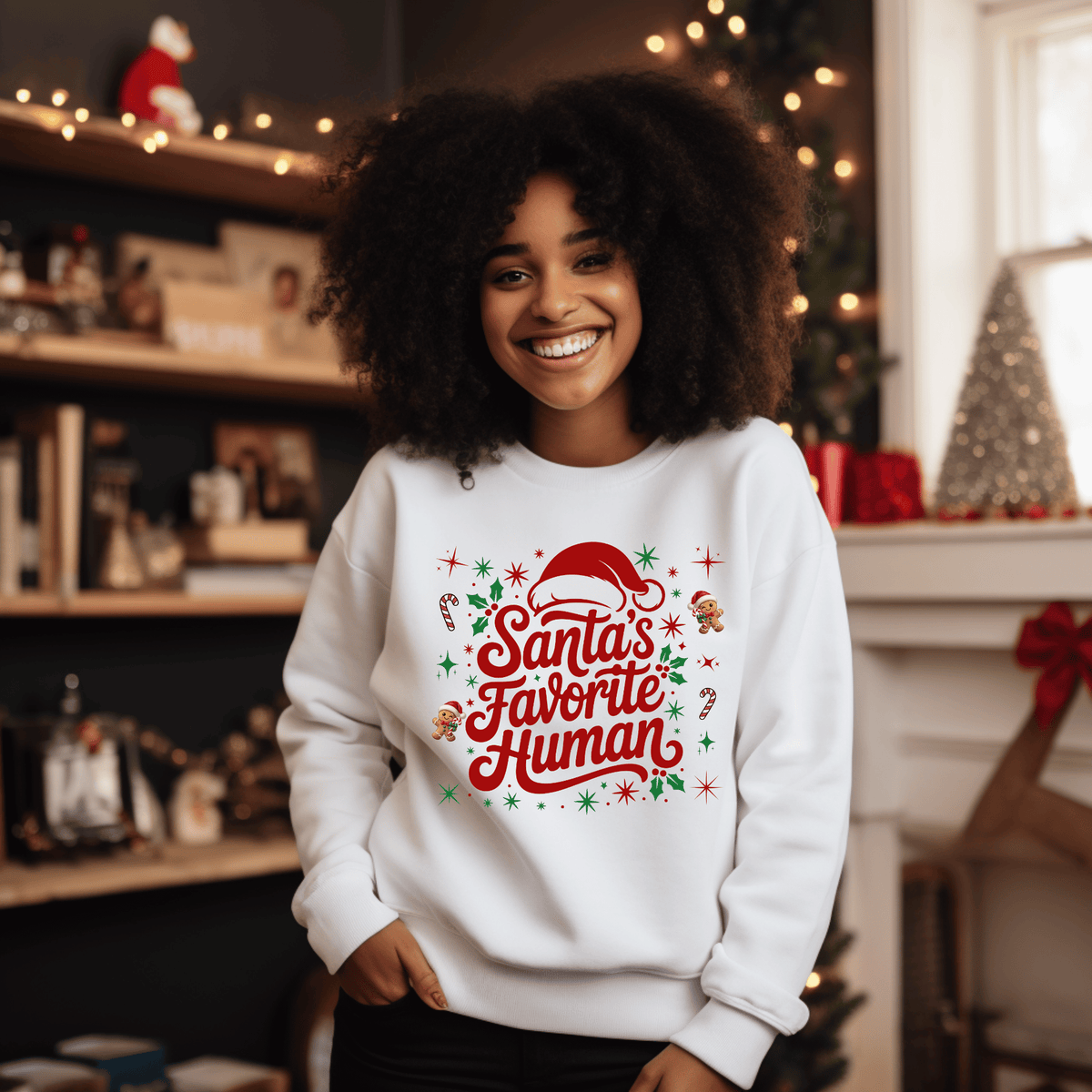 Folded sand crewneck sweatshirt with 'Santa's Favorite Human' red lettering, Santa hat, and gingerbread graphics on a neutral wooden background.