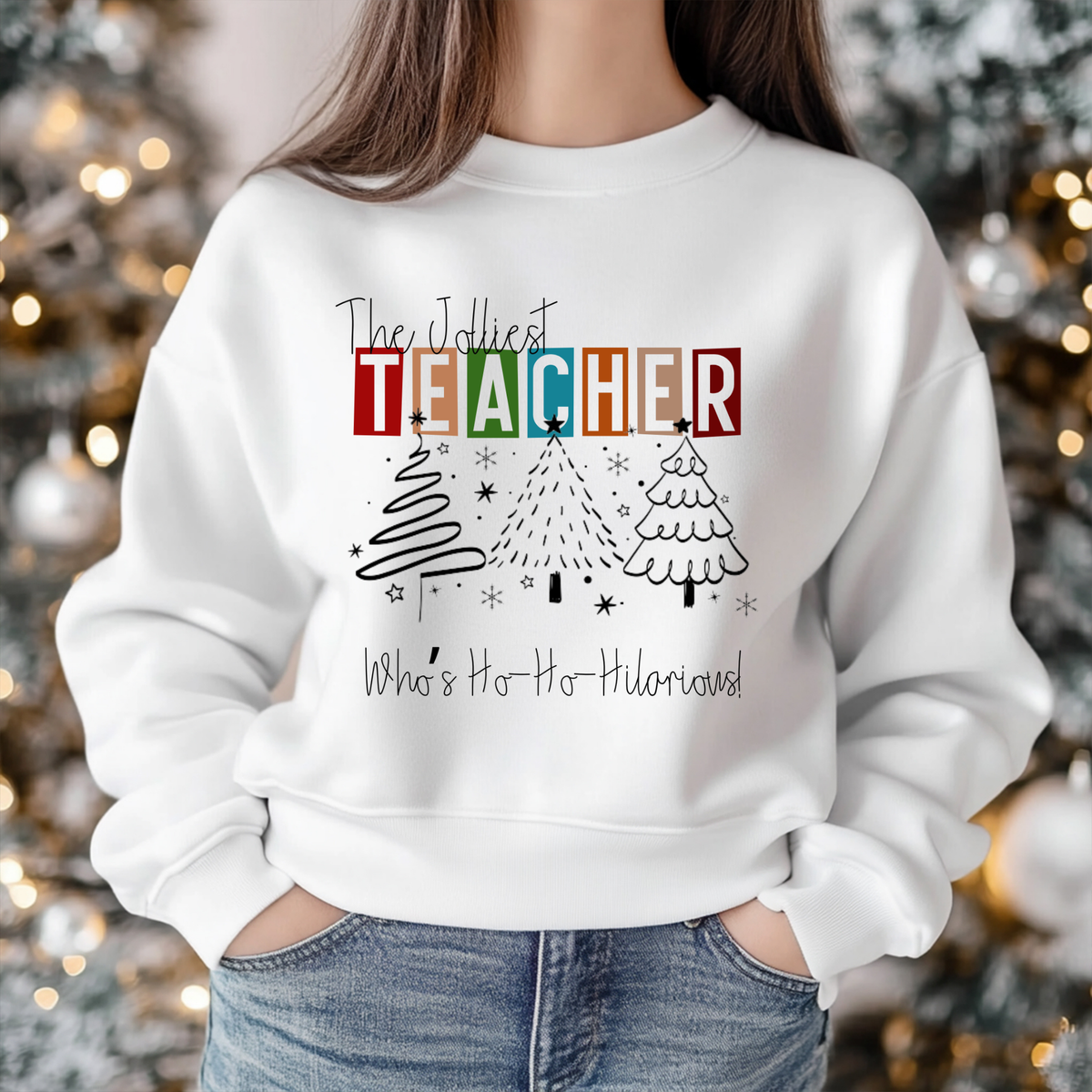 A person wearing a white crewneck sweatshirt with a festive Christmas graphic that reads 'The Jolliest TEACHER Who's Ho-Ho-Humorist!' above three hand-drawn Christmas trees, shown against a blurred background of a decorated Christmas tree with warm lights.