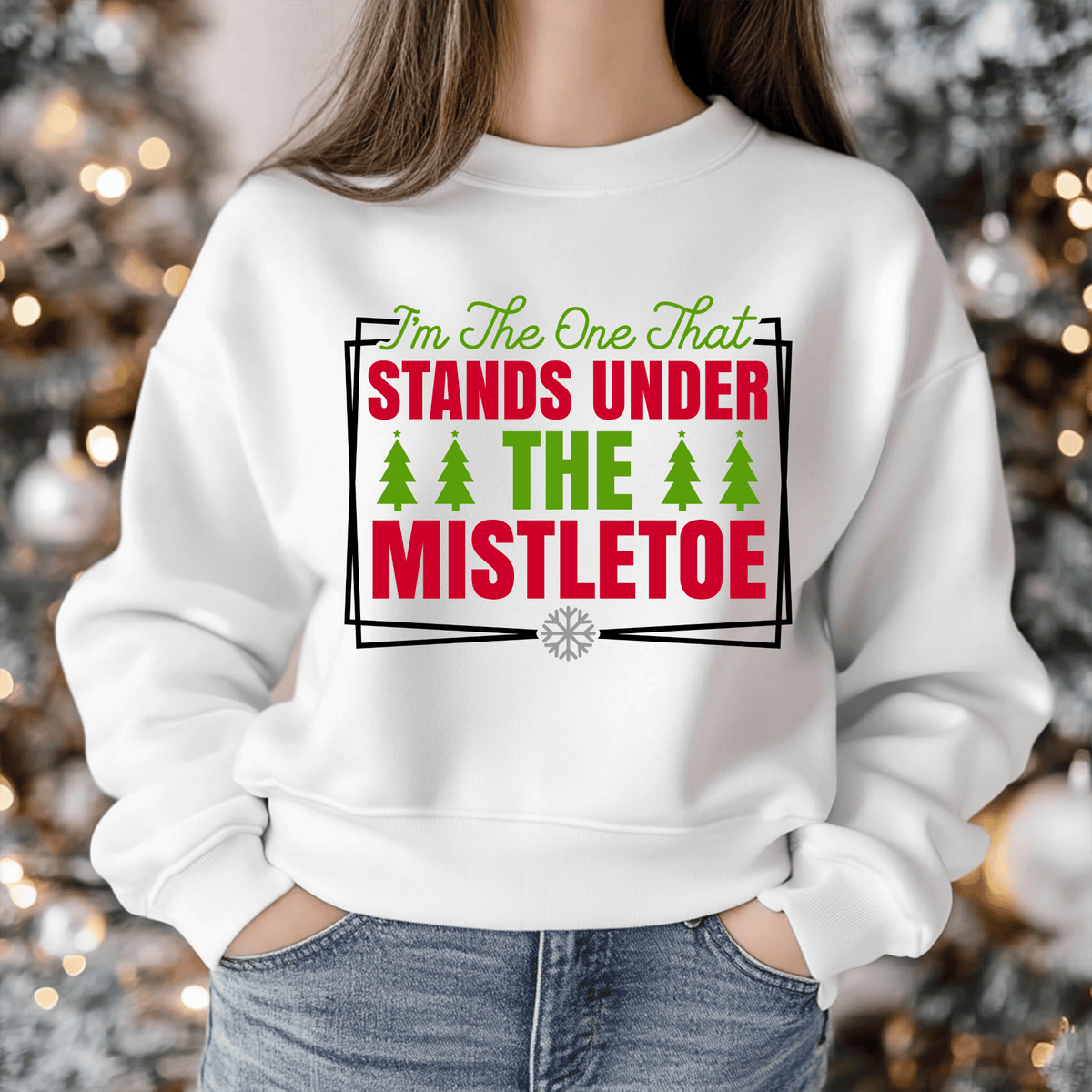 A person wearing a white crewneck sweatshirt with a festive Christmas graphic that reads 'I'm The One That STANDS UNDER THE MISTLETOE' in red and green text with green pine trees, set against a blurred holiday tree background.