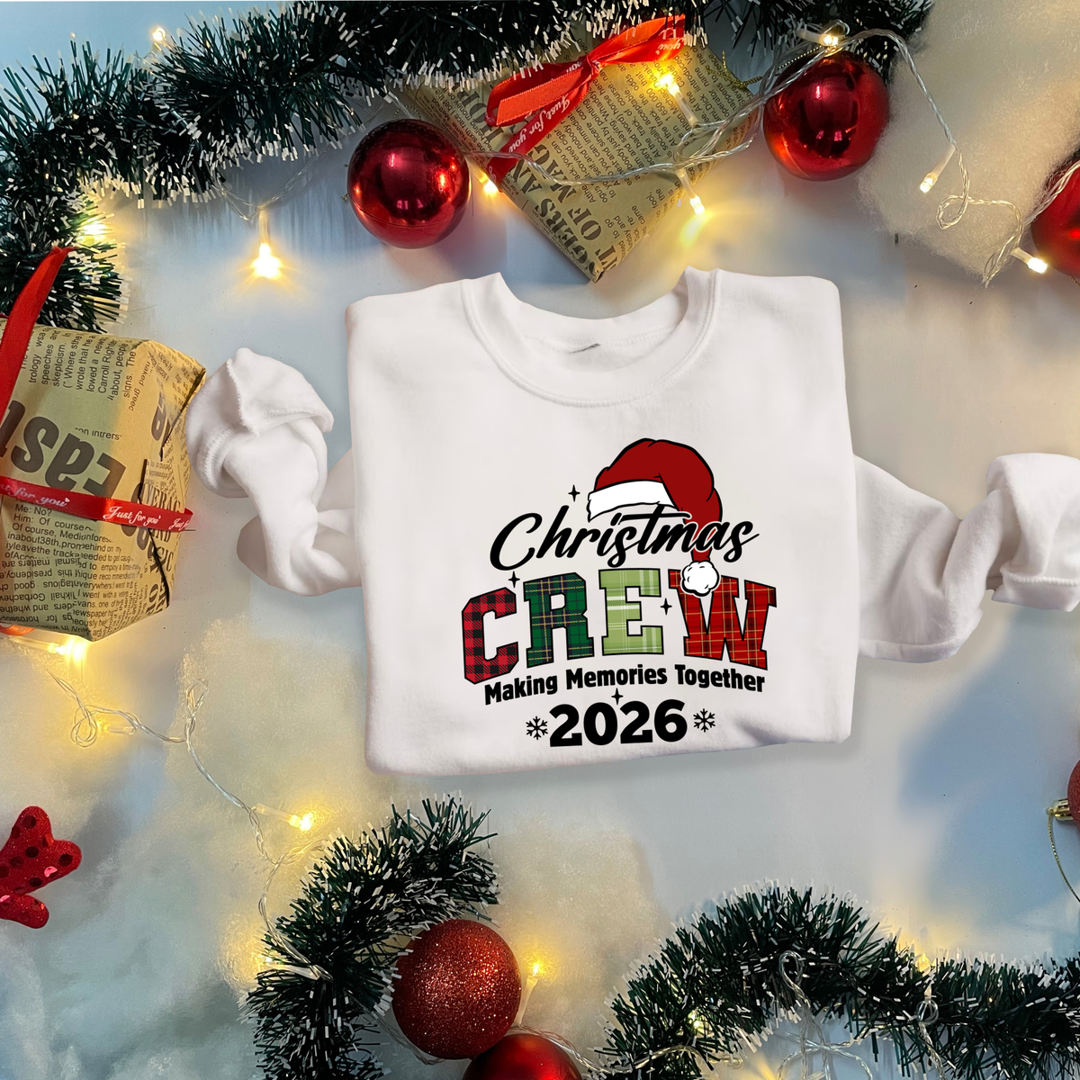 A white long-sleeve crewneck sweatshirt with a festive Christmas graphic reading 'Christmas CREW Making Memories Together 2026' in red, green, and black lettering with a Santa hat, presented in a holiday flat-lay setting with lights, ornaments, and wrapped gifts.