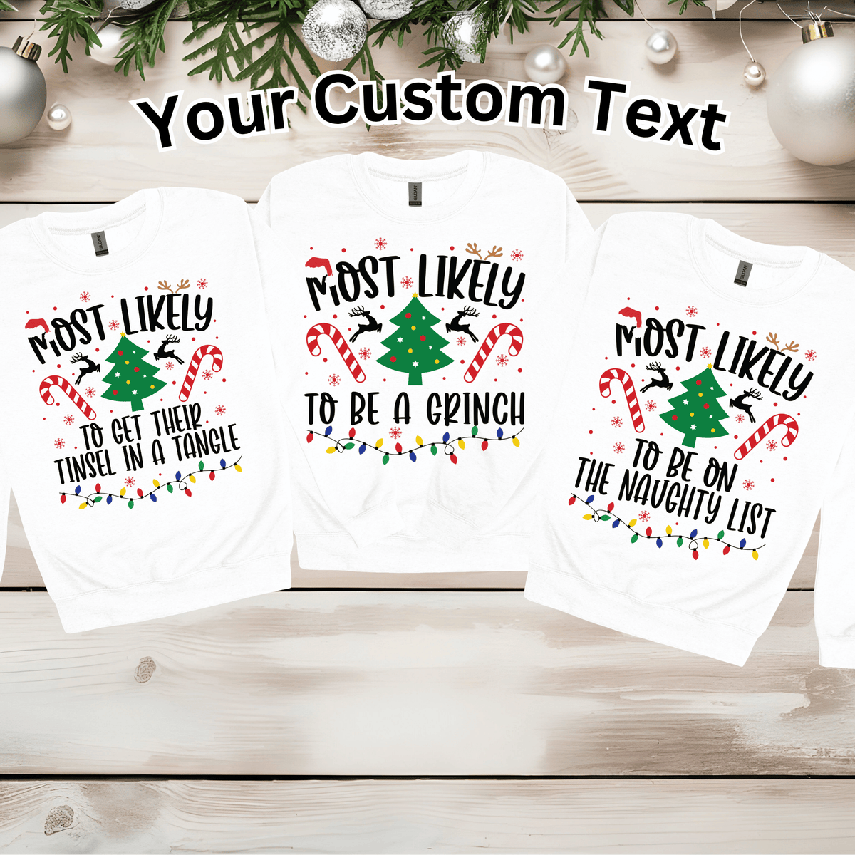 Three white long-sleeve t-shirts with a custom Christmas graphic print, displayed on a light wood background with holiday decorations. The graphic features a green Christmas tree, candy canes, Santa silhouettes, and colorful Christmas lights, with the text 'Most Likely To Be A Grinch!' and 'Most Likely To Be On The Naughty List'.