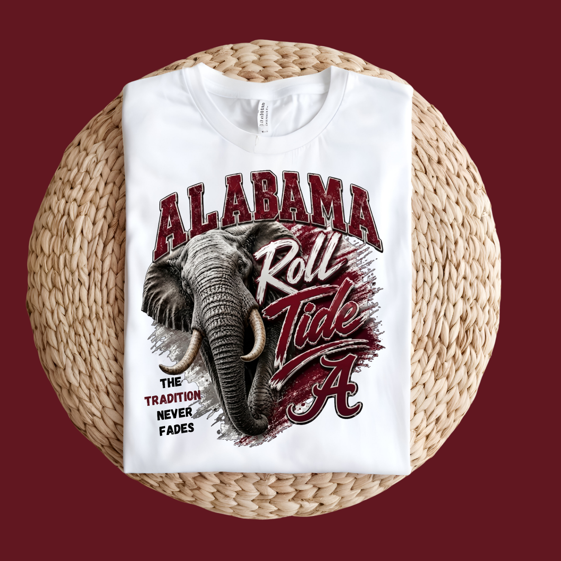 A folded white graphic T-shirt displayed on a round woven straw mat against a dark red background. The shirt features a large graphic with an elephant, the text 'ALABAMA Roll Tide', and 'THE TRADITION NEVER FADES'.