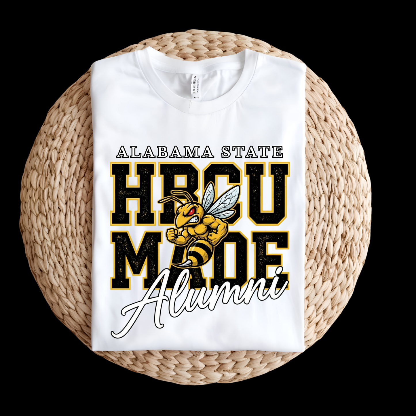 A white folded T-shirt with a large Alabama State HBCU Made Alumni graphic, displayed on a round woven straw mat against a black background.