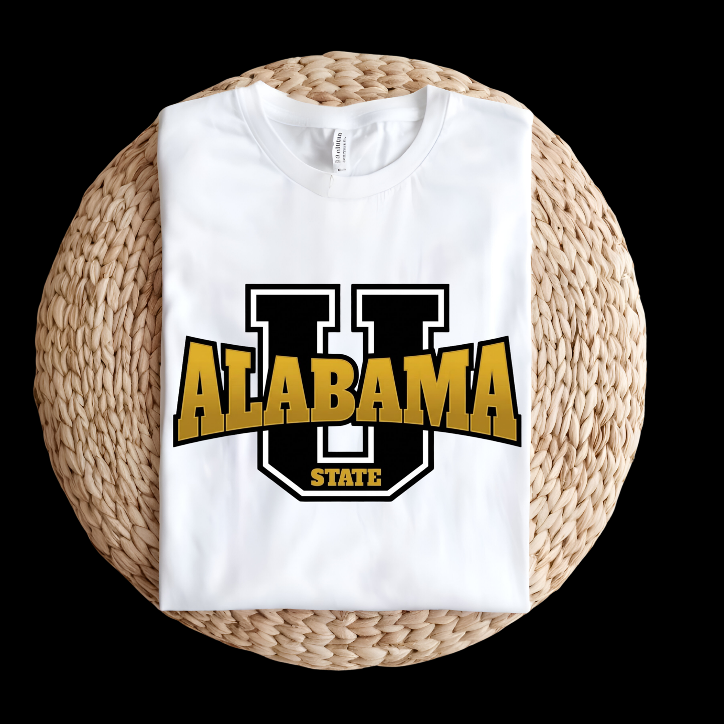 A folded white t-shirt featuring a large graphic of a black and gold 'U' with 'ALABAMA STATE' printed across it, displayed on a round woven straw mat against a solid black background.