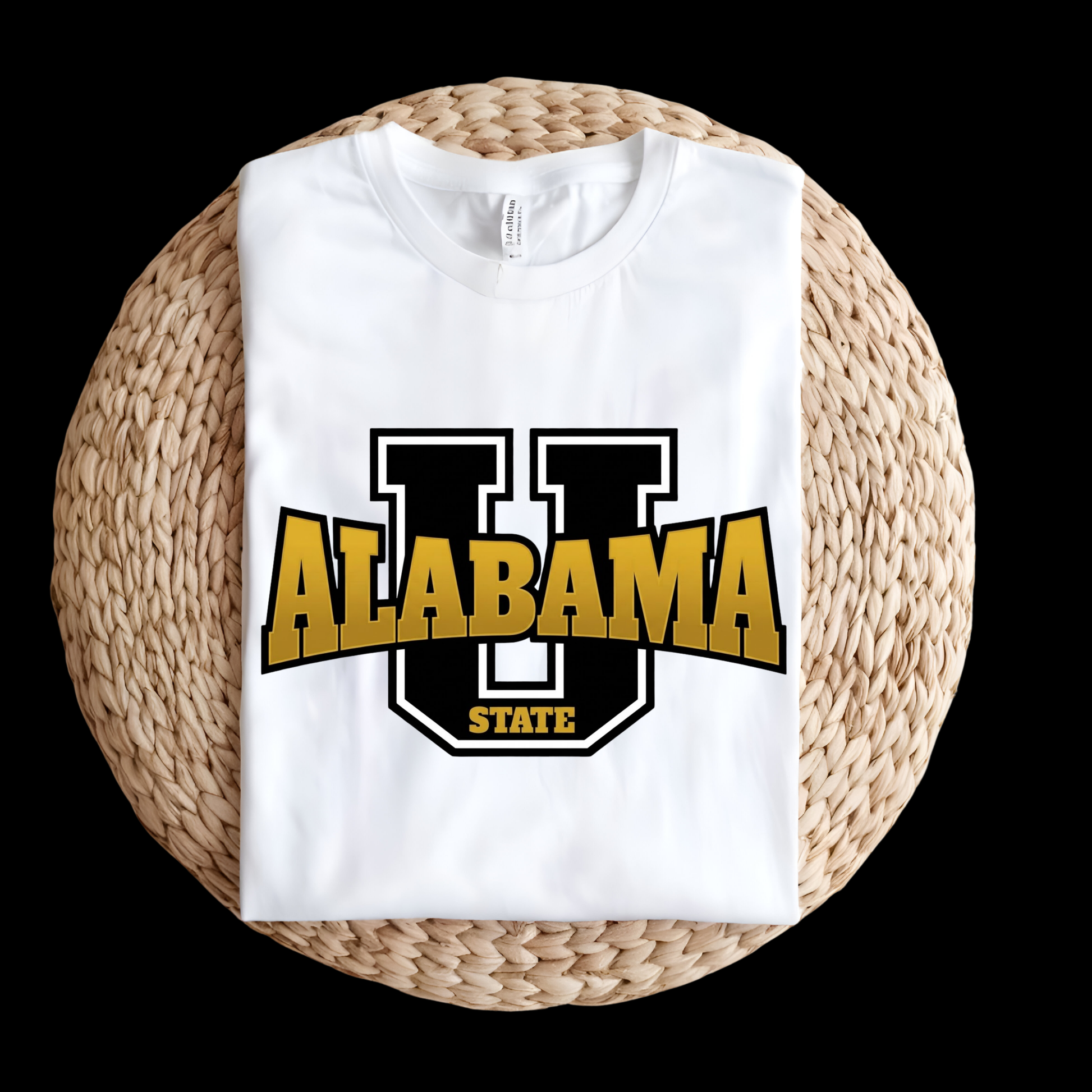 A folded white t-shirt featuring a large graphic of a black and gold 'U' with 'ALABAMA STATE' printed across it, displayed on a round woven straw mat against a solid black background.