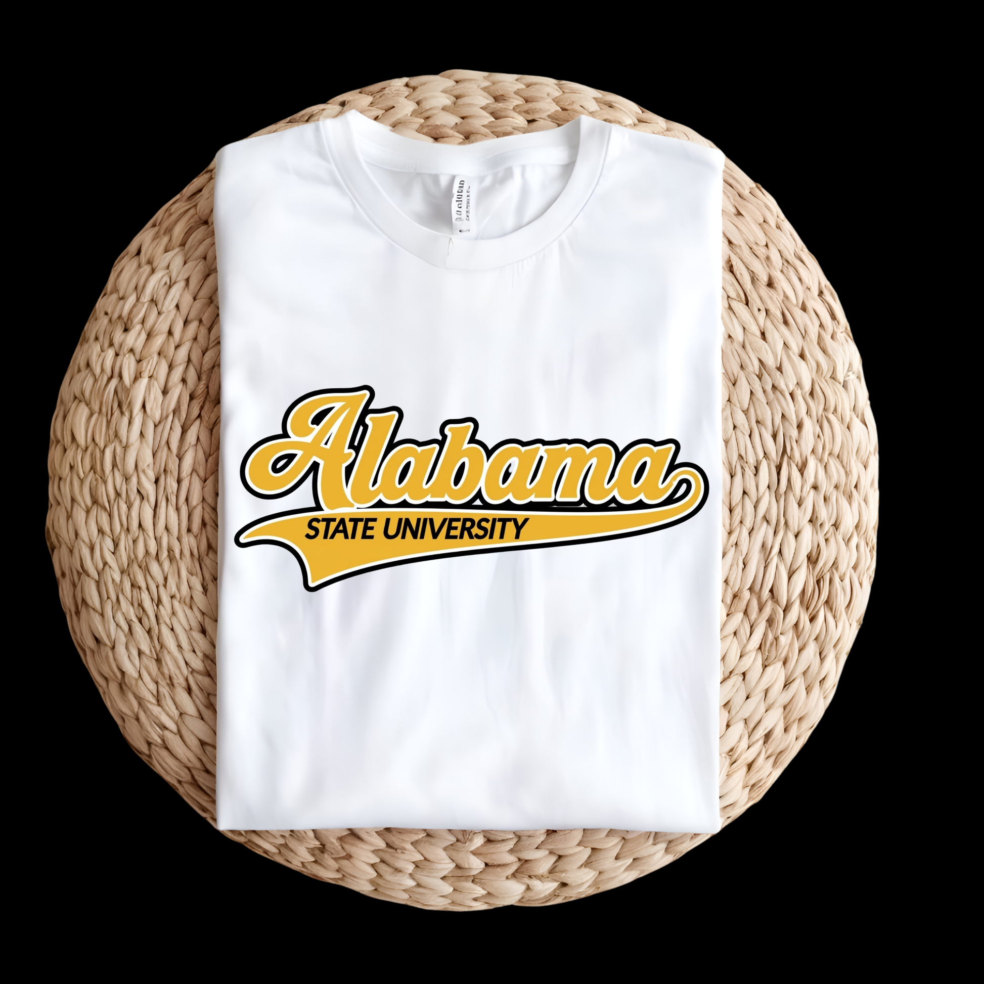 A folded white t-shirt with a yellow and black 'Alabama State University' graphic, displayed on a round woven straw mat against a solid black background.