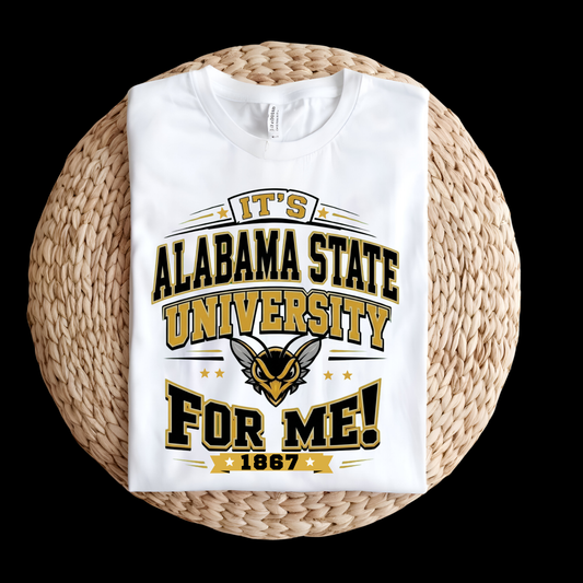 A white short-sleeve T-shirt folded and displayed on a round woven straw mat against a solid black background. The shirt features a bold graphic print in black and gold reading 'IT'S ALABAMA STATE UNIVERSITY FOR ME!' with '1867' at the bottom and an Alabama State Hornets mascot logo in the center.