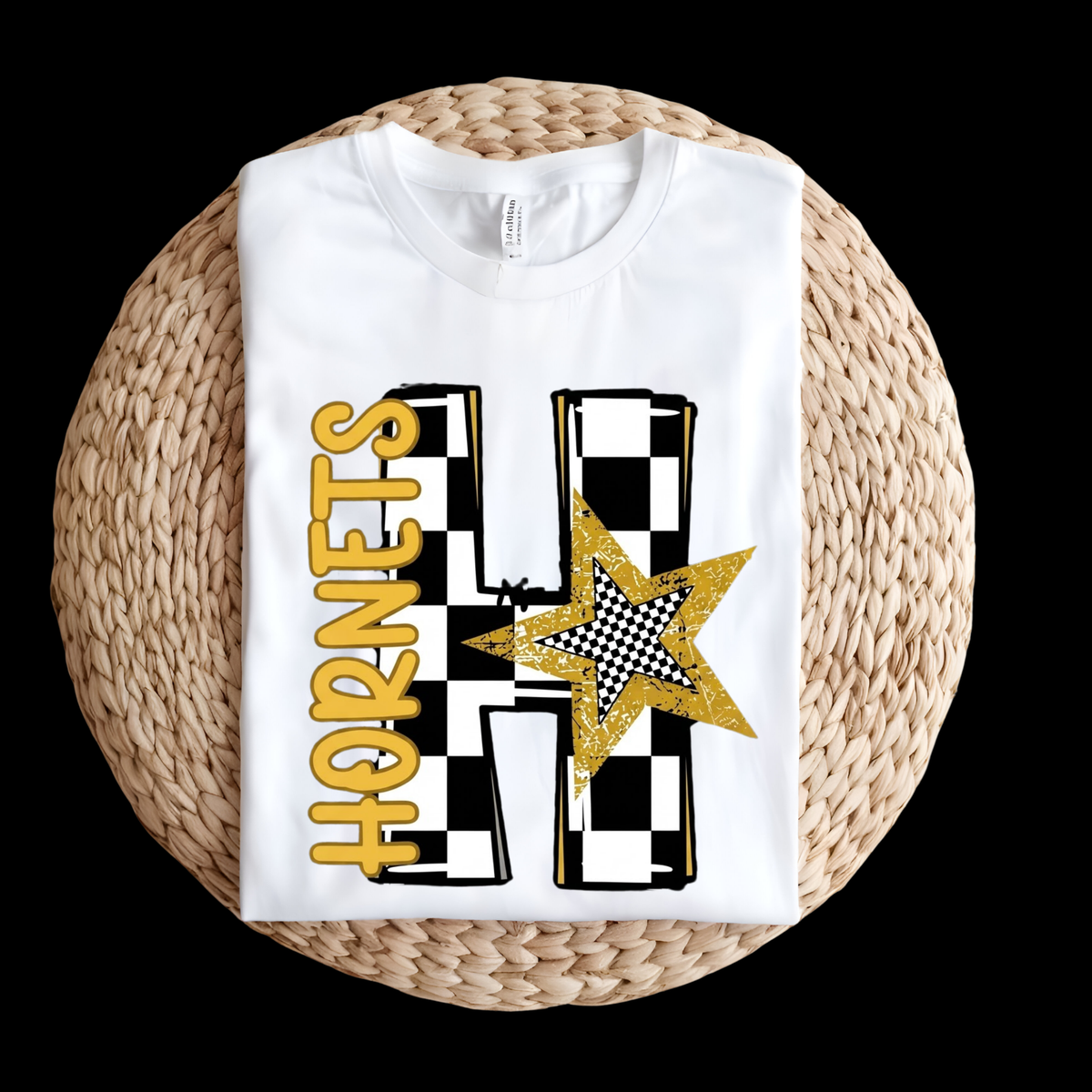 A folded white t-shirt with a black and gold checkerboard 'H' and the word 'HORNETS' in gold, displayed on a round woven straw mat against a black background.