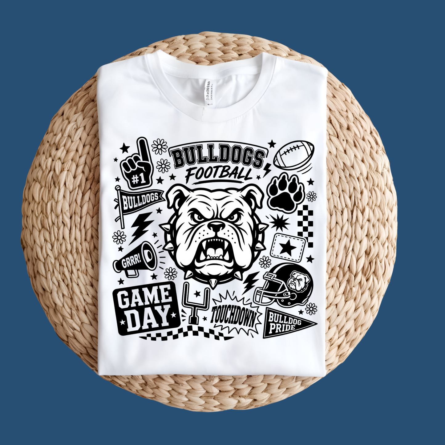 A white short-sleeve T-shirt laid flat on a round woven straw mat against a black background. The shirt has a large front graphic reading “WE ARE Bulldogs” in distressed red, black, and white lettering.