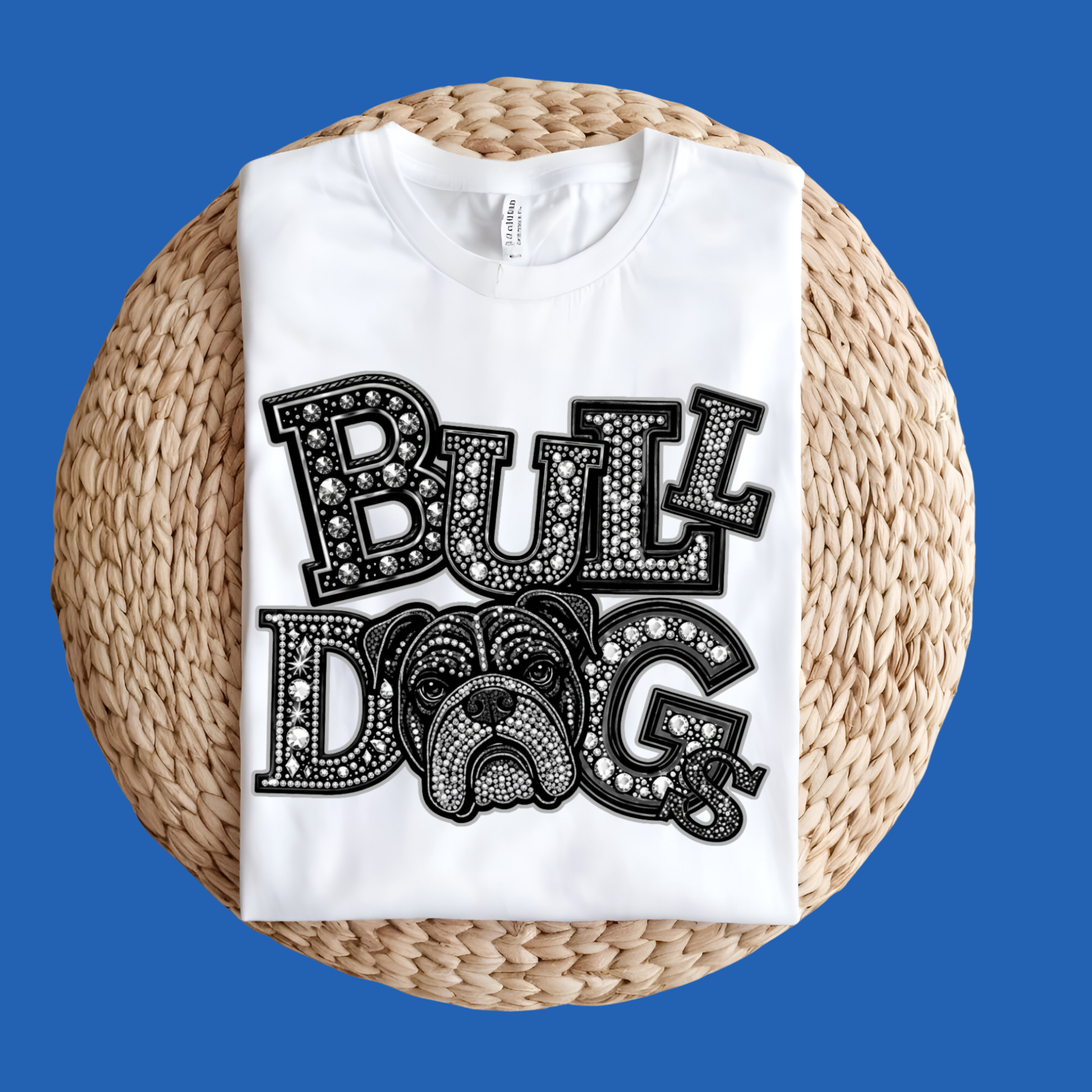 A white short-sleeve T-shirt laid flat on a round woven straw mat against a black background. The shirt has a large front graphic reading “WE ARE Bulldogs” in distressed red, black, and white lettering.