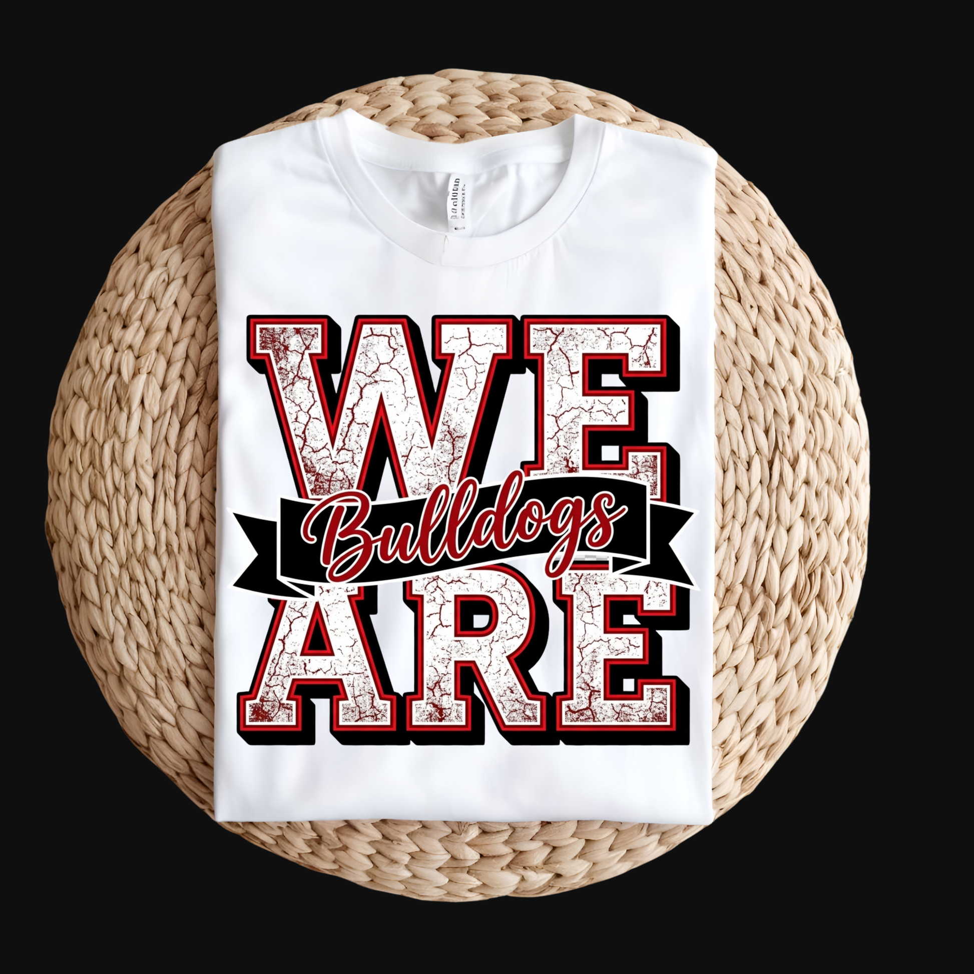 A white short-sleeve T-shirt laid flat on a round woven straw mat against a black background. The shirt has a large front graphic reading “WE ARE Bulldogs” in distressed red, black, and white lettering.