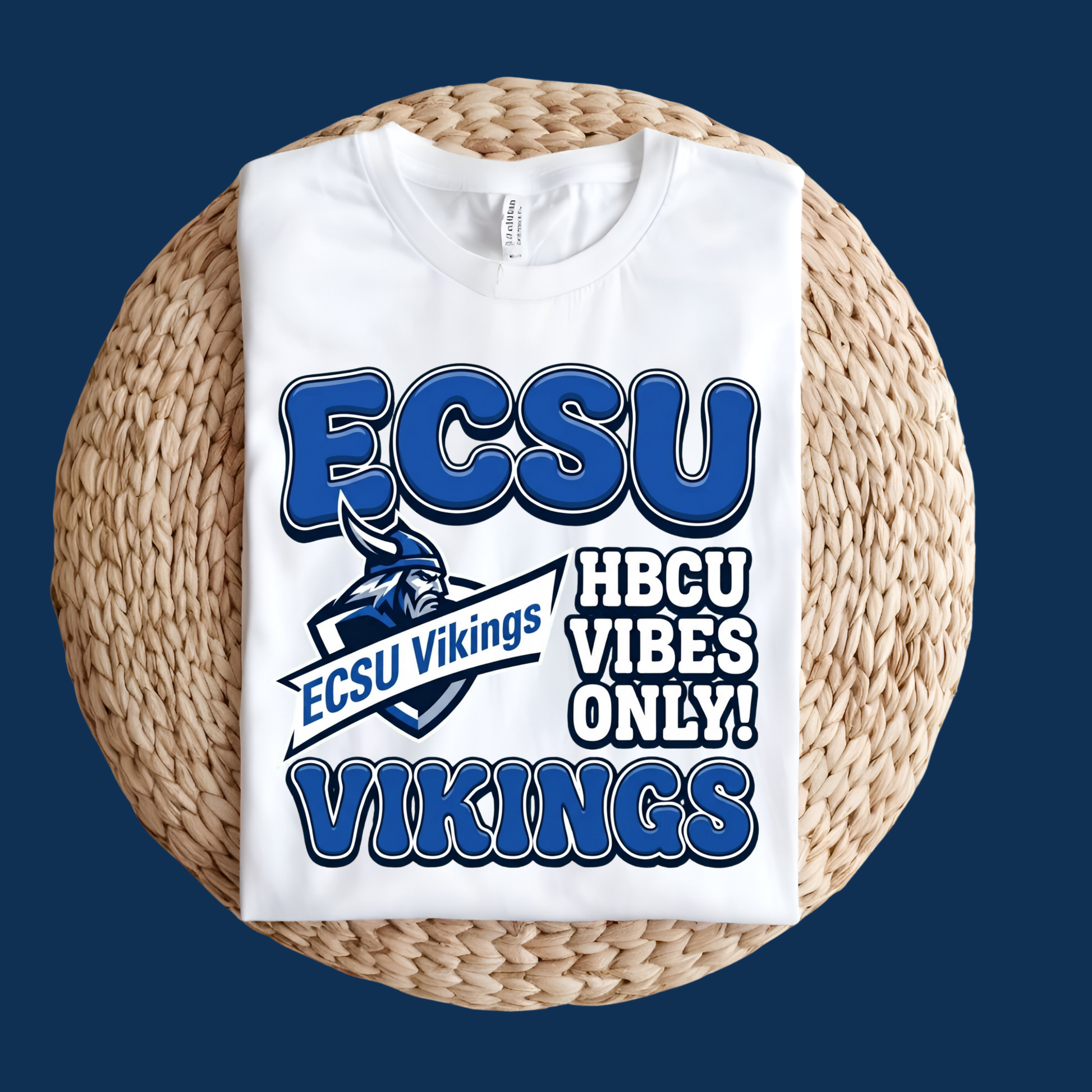A white crewneck t-shirt with a blue and white 'ECSU Vikings' graphic, displayed flat on a round woven straw mat against a dark blue background.