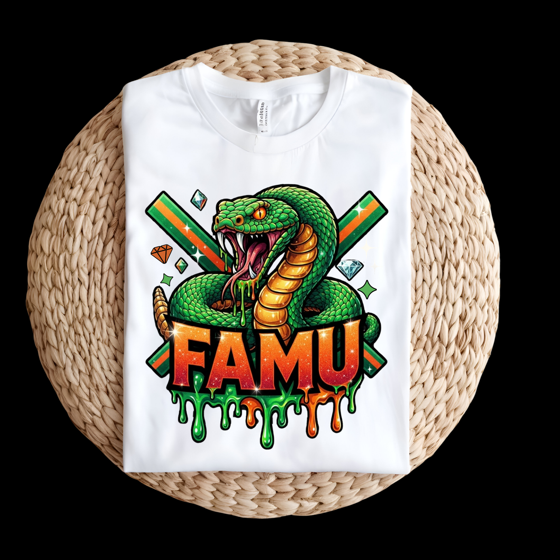 A white short-sleeve T-shirt laid flat on a round woven straw mat, featuring a large colorful graphic of a green snake with crossed green and orange swords, diamonds, and the word "FAMU" in bold orange-red letters with dripping green and orange paint details.