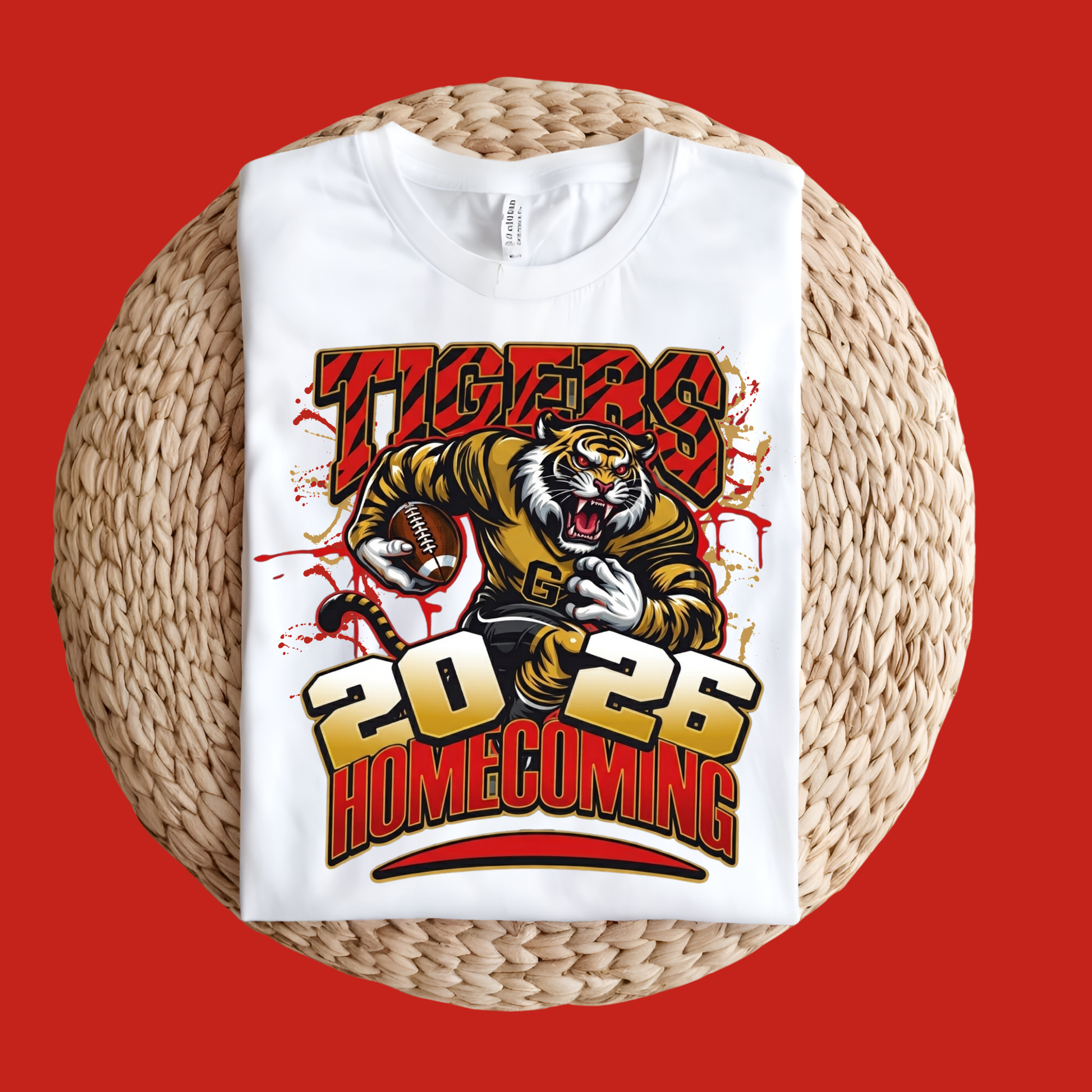 A white short-sleeve T-shirt laid flat on a round woven straw mat against a solid red background. The shirt features a large graphic print with the word 'TIGERS' in red, a roaring tiger mascot holding a football, and '2026 HOMECOMING' in bold yellow and red lettering.