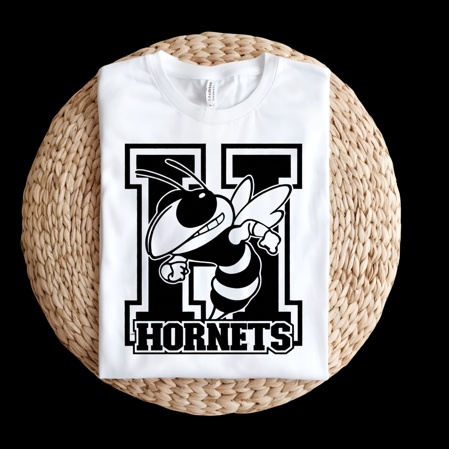 A folded white t-shirt with a black and white 'N Hornets' graphic featuring a hornet mascot, displayed on a round woven straw mat against a solid black background.