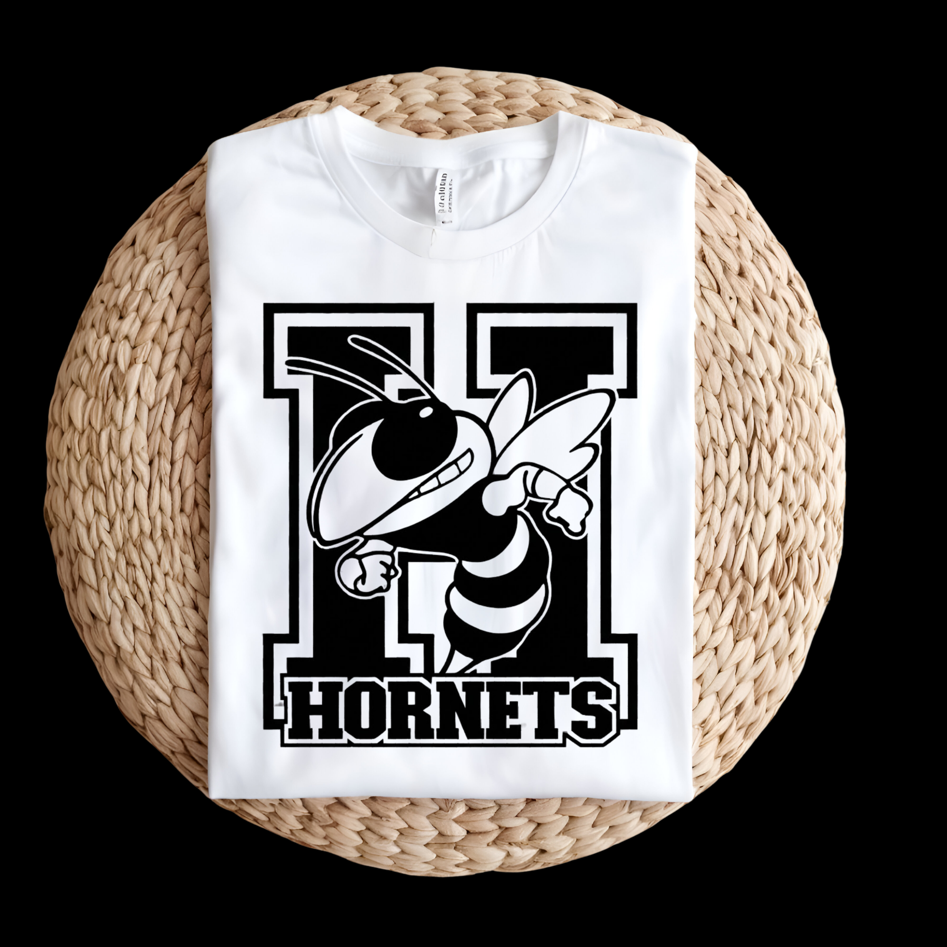 A folded white t-shirt with a black and white 'N Hornets' graphic featuring a hornet mascot, displayed on a round woven straw mat against a solid black background.