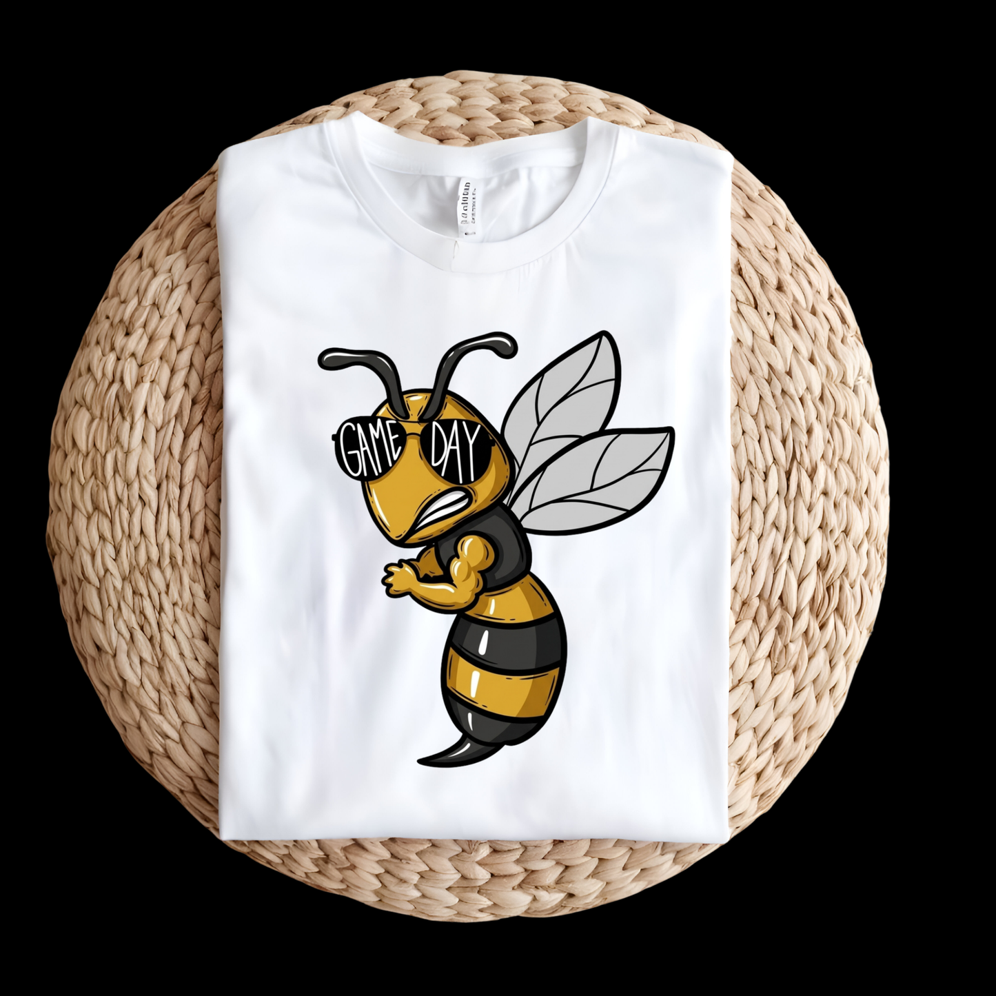 A folded white t-shirt displayed on a round woven straw mat against a solid black background. The t-shirt features a cartoon graphic of a bee wearing black and yellow stripes and sunglasses with the text 'GAME DAY' written on them.