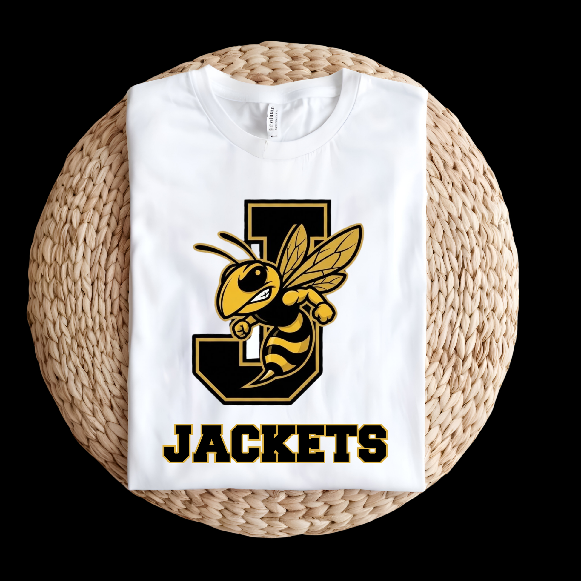 A folded white t-shirt with a black and yellow 'J' jacket mascot and the word 'JACKETS' printed on the front, displayed on a round woven straw mat against a black background.