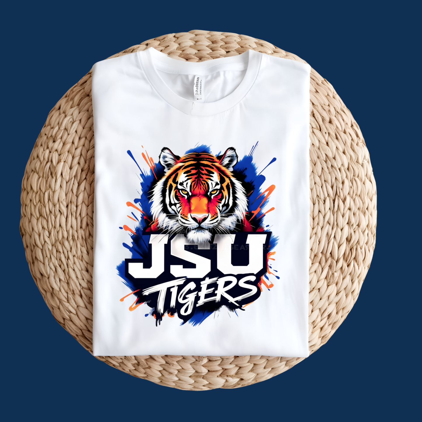 A white crewneck t-shirt folded flat on a round woven straw mat, featuring a graphic of a tiger's face with a red nose and blue paint splatters, above the text 'JSU TIGERS' in white and black.