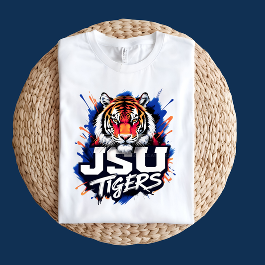 A white crewneck t-shirt folded flat on a round woven straw mat, featuring a graphic of a tiger's face with a red nose and blue paint splatters, above the text 'JSU TIGERS' in white and black.