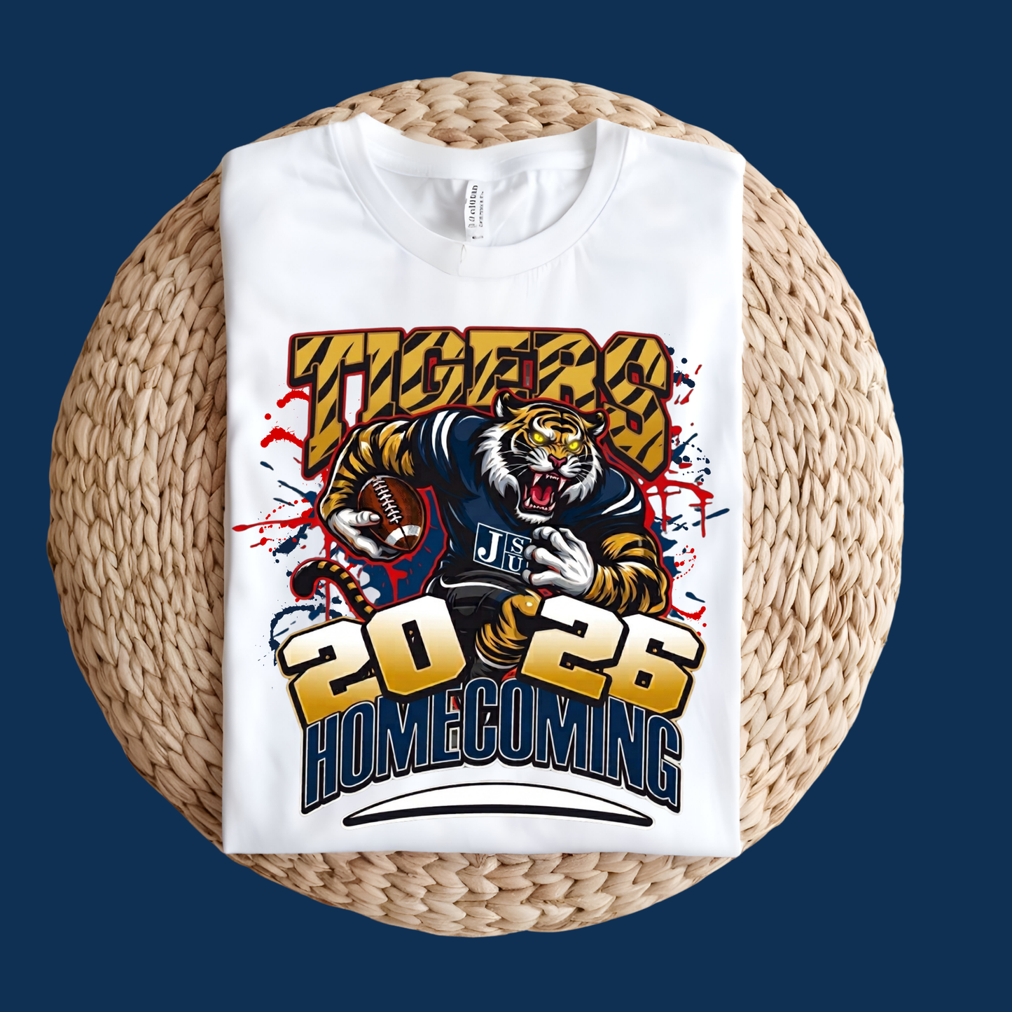 A white short-sleeve T-shirt laid flat on a round woven straw mat, featuring a large football-themed graphic with a tiger, the word 'Tigers', and '2026 Homecoming' in bold yellow, blue, red, and black lettering.