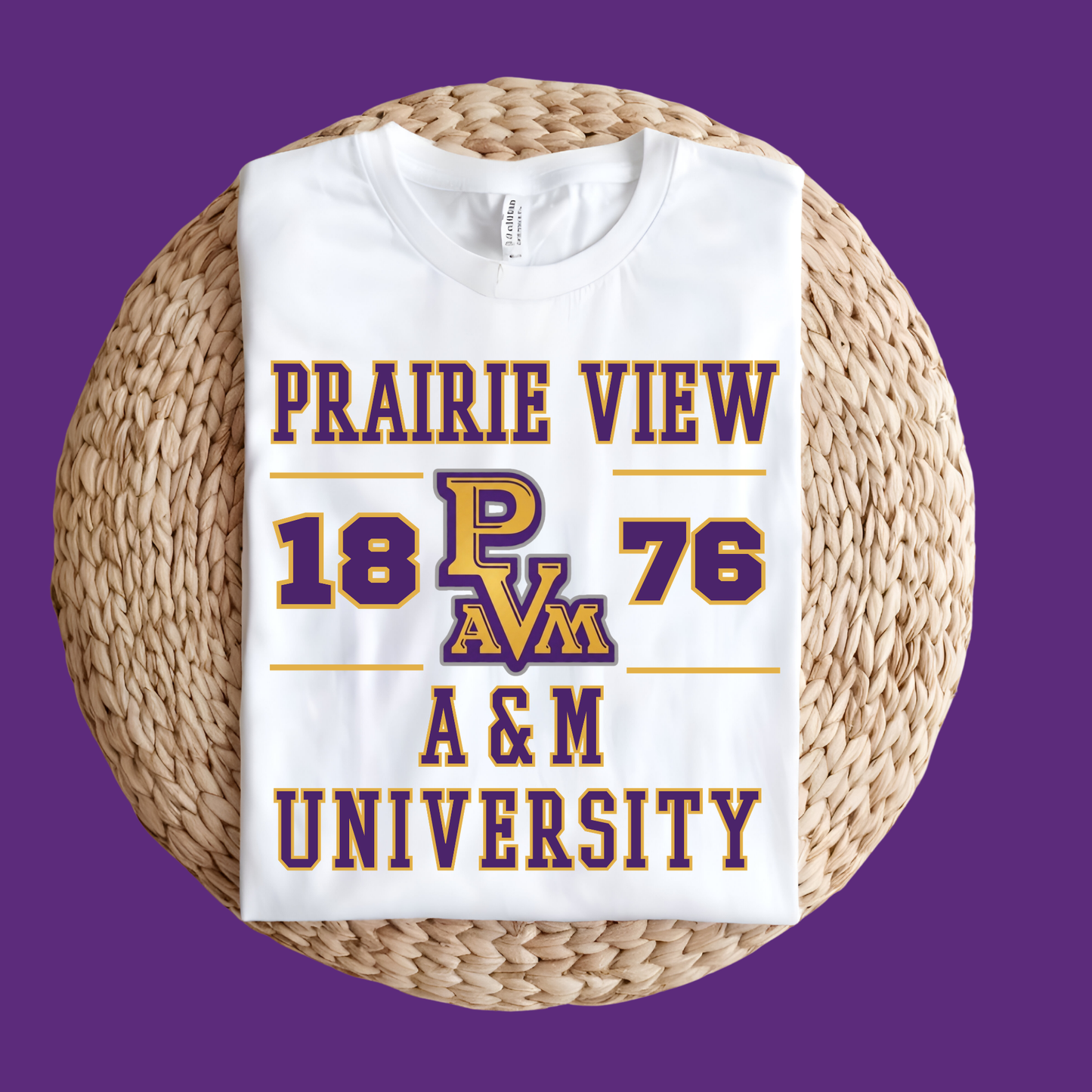A folded white t-shirt with a purple and gold 'Prairie View 1876 A&M University' graphic, displayed on a round woven straw mat against a solid purple background.