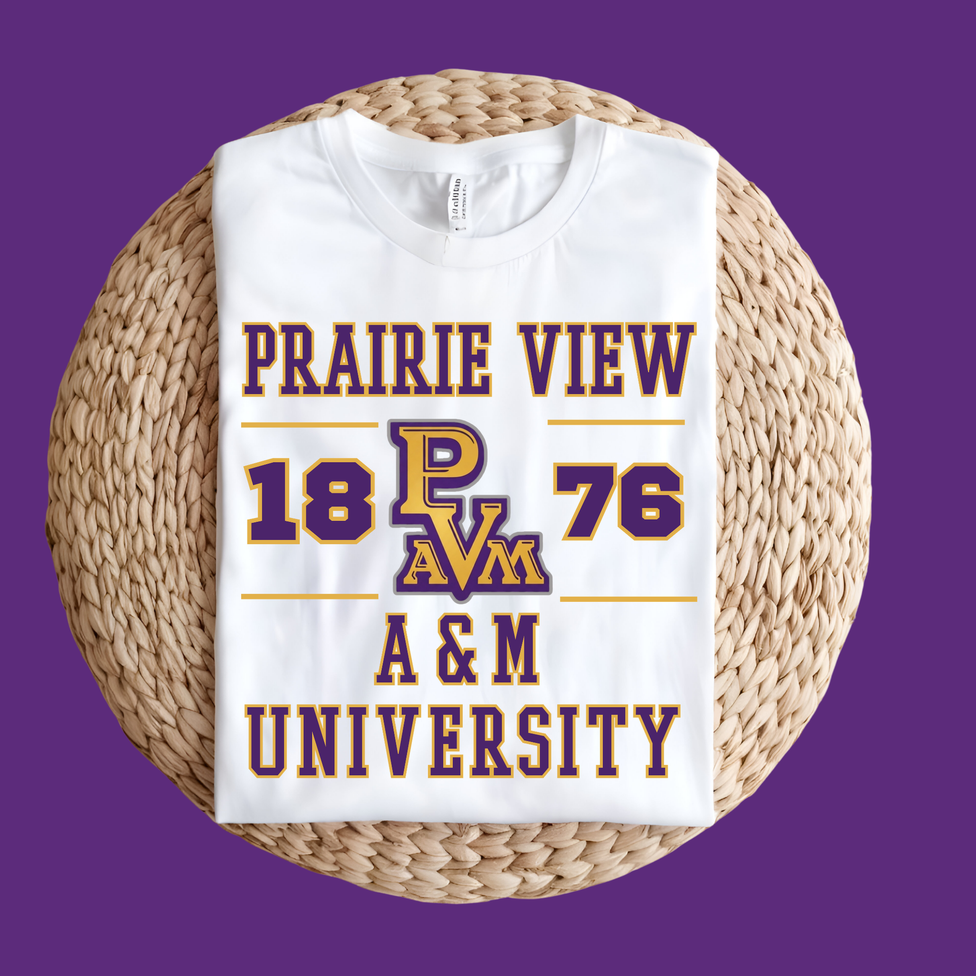 A folded white t-shirt with a purple and gold 'Prairie View 1876 A&M University' graphic, displayed on a round woven straw mat against a solid purple background.