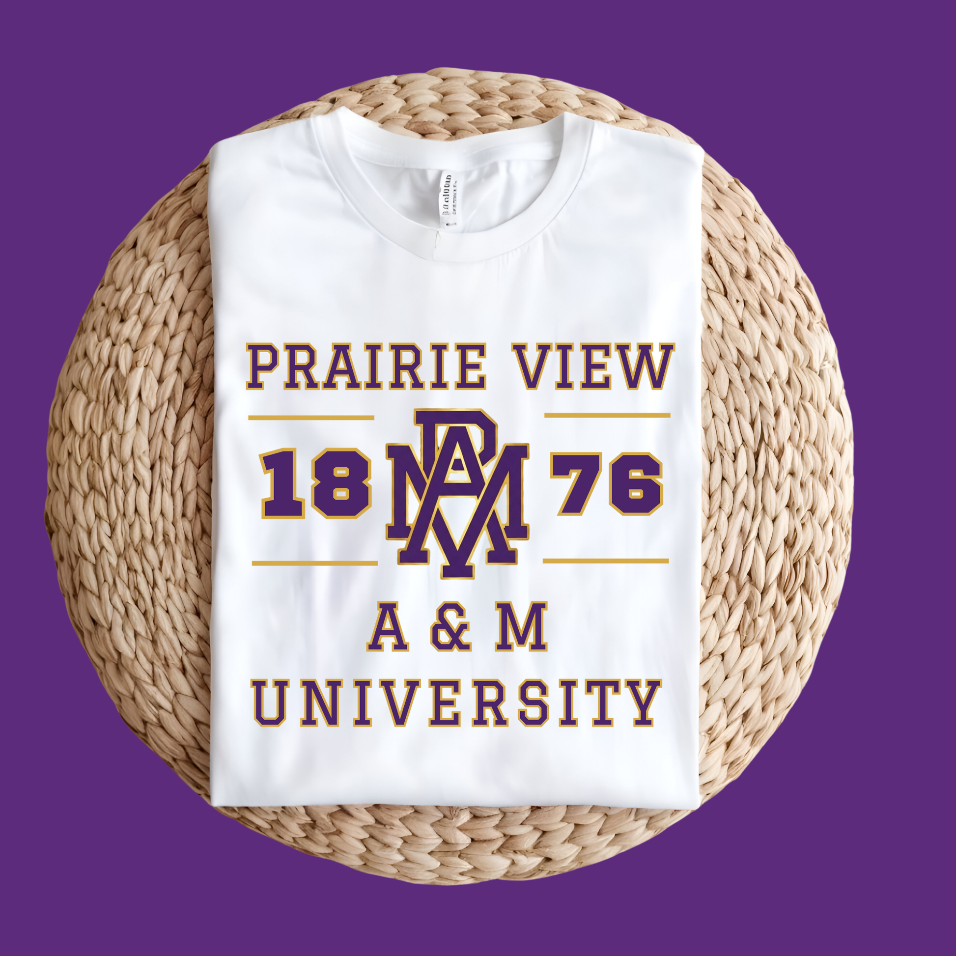 A folded white t-shirt with a purple and gold graphic reading 'PRAIRIE VIEW 1876 A & M UNIVERSITY' and an 'AM' monogram, displayed on a round woven straw mat against a solid purple background.