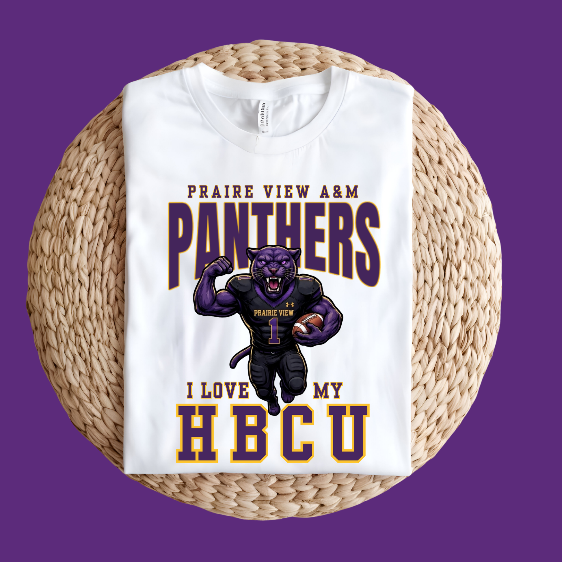 A white short-sleeve T-shirt laid flat on a round woven straw mat against a solid purple background. The shirt features a graphic print with the text 'PRAIRIE VIEW A&M PANTHERS I LOVE MY HBCU' and an illustration of a purple panther mascot wearing a football uniform and holding a football.