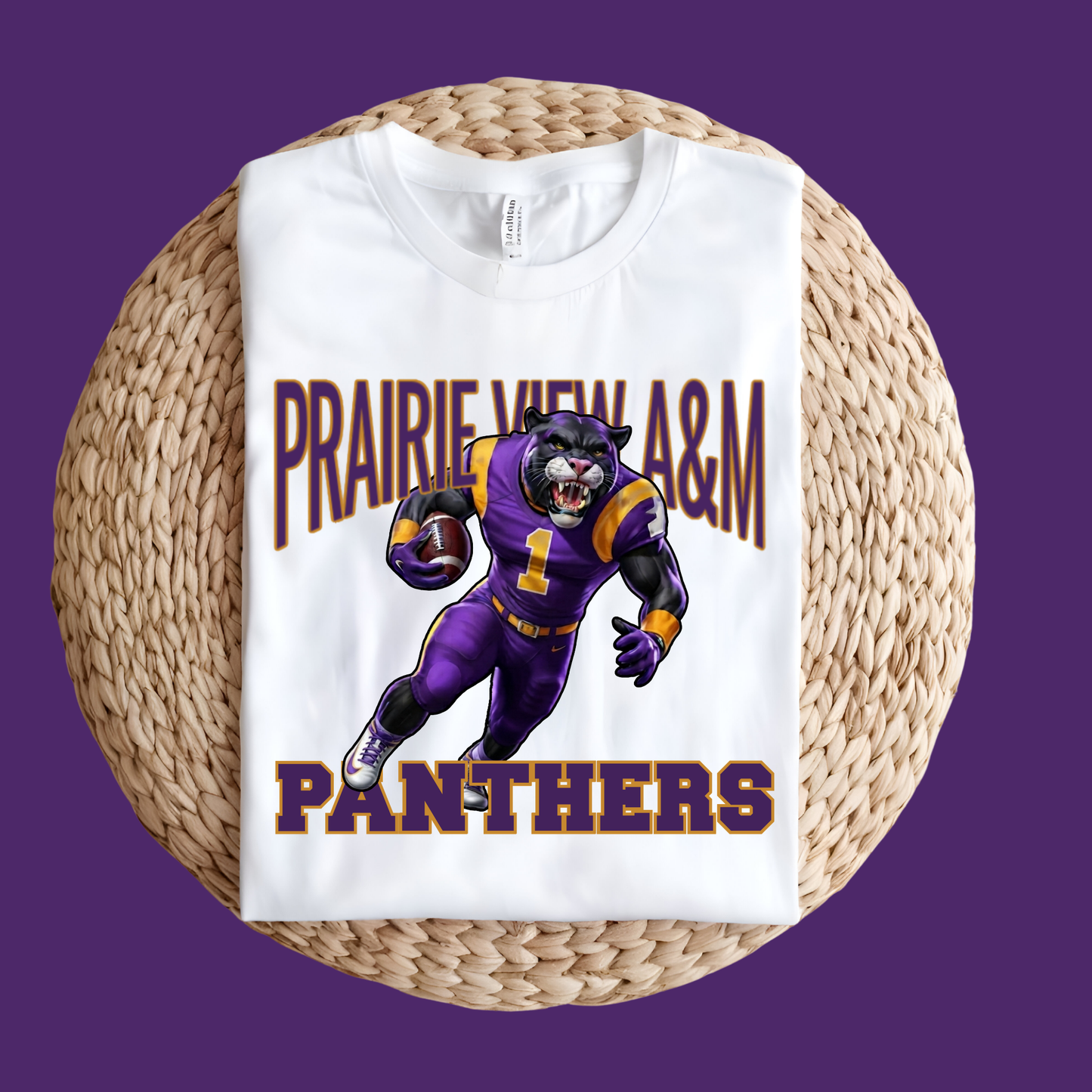 A white short-sleeve T-shirt laid flat on a round woven straw mat, featuring a large graphic of a purple and gold football player with the text 'PRAIRIE VIEW A&M PANTHERS' on the front, set against a solid purple background.