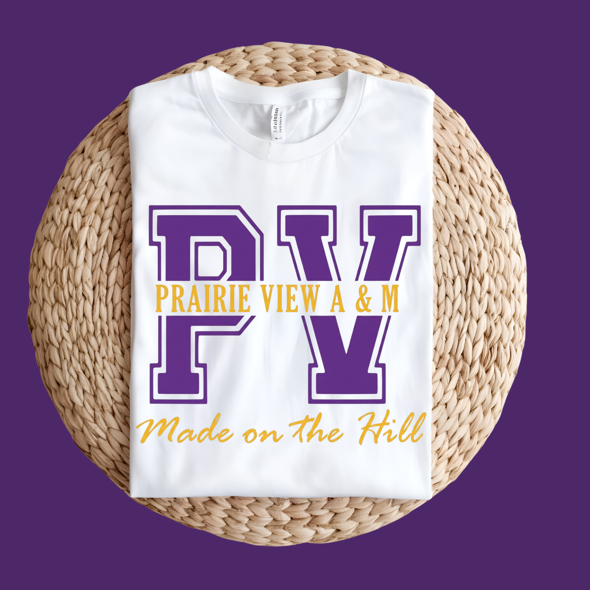 A white short-sleeve T-shirt laid flat on a round woven straw mat, featuring a large graphic of a purple and gold football player with the text 'PRAIRIE VIEW A&M PANTHERS' on the front. The shirt is photographed against a solid purple background.
