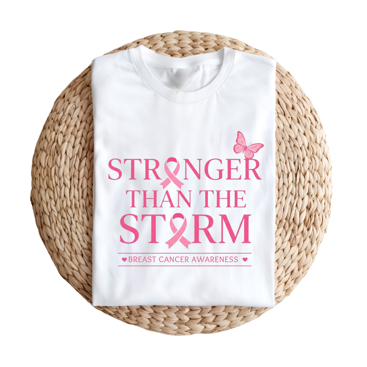 A folded white T-shirt with pink text reading 'STRONGER THAN THE STORM' and 'BREAST CANCER AWARENESS' with a pink ribbon and butterfly, displayed on a round woven straw mat against a white background.