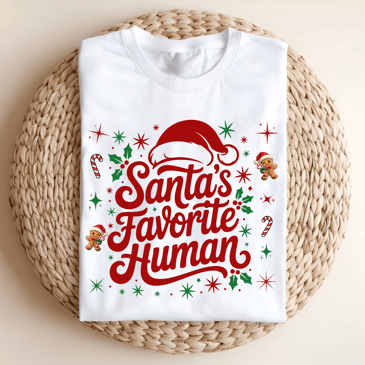 Folded sand crewneck sweatshirt with 'Santa's Favorite Human' red lettering, Santa hat, and gingerbread graphics on a neutral wooden background.