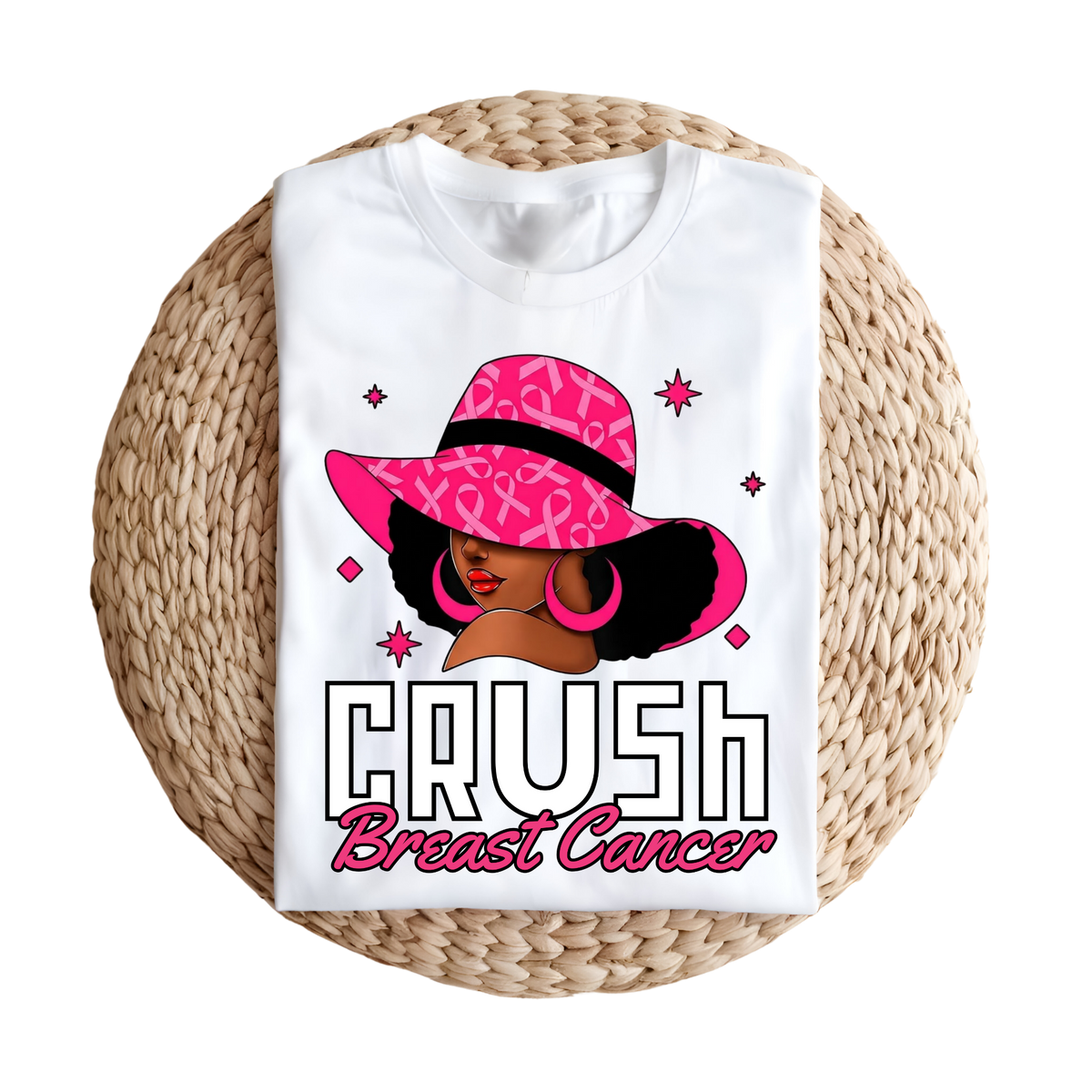 A white short-sleeve T-shirt laid flat on a round woven straw mat, featuring a colorful graphic of a woman in a pink ribbon-patterned hat with the text 'CRUSH Breast Cancer'.