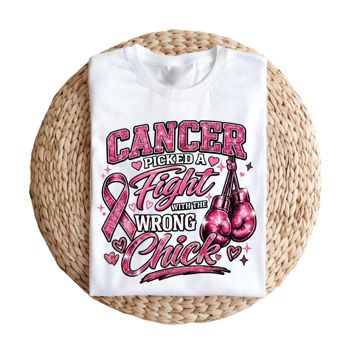 A white short-sleeve T-shirt laid flat on a round woven straw mat, featuring a large pink and black graphic that reads “I AM A SURVIVOR” with pink ribbons, butterflies, and hearts.