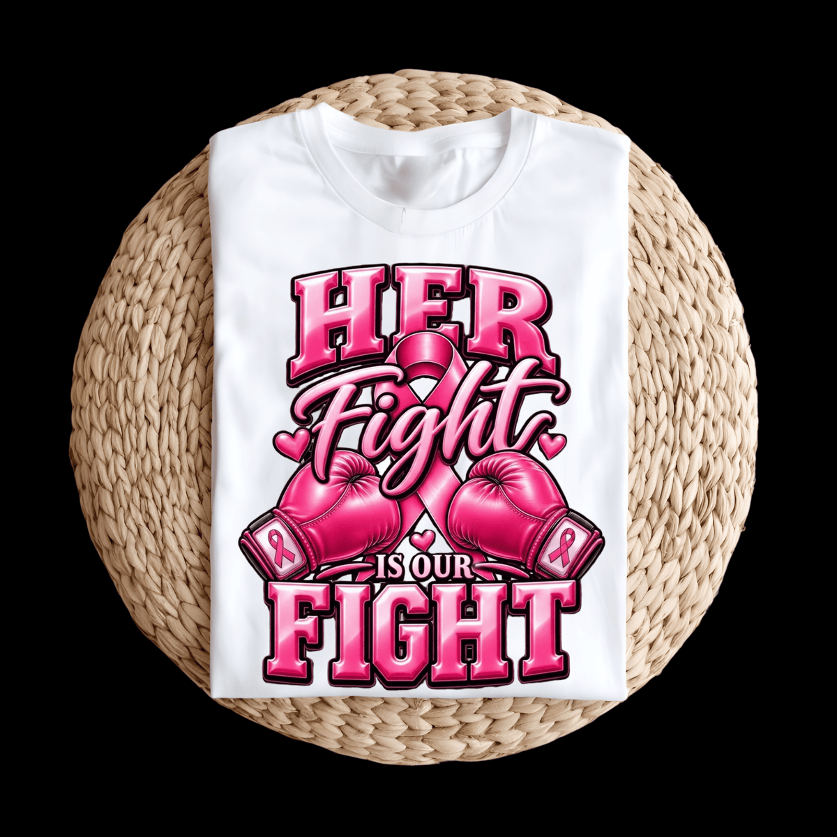 A folded white T-shirt displayed on a round woven straw mat against a black background. The shirt features a bold pink graphic with the words 'HER Fight IS OUR FIGHT' and pink awareness ribbons and boxing gloves.