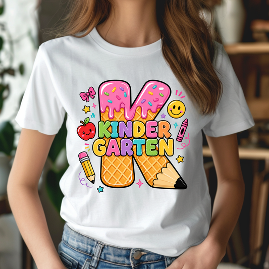 A woman wearing a white graphic t-shirt with a colorful 'Kinder Garten' design featuring two ice cream cones shaped like pencils, a smiling apple, and school supplies.