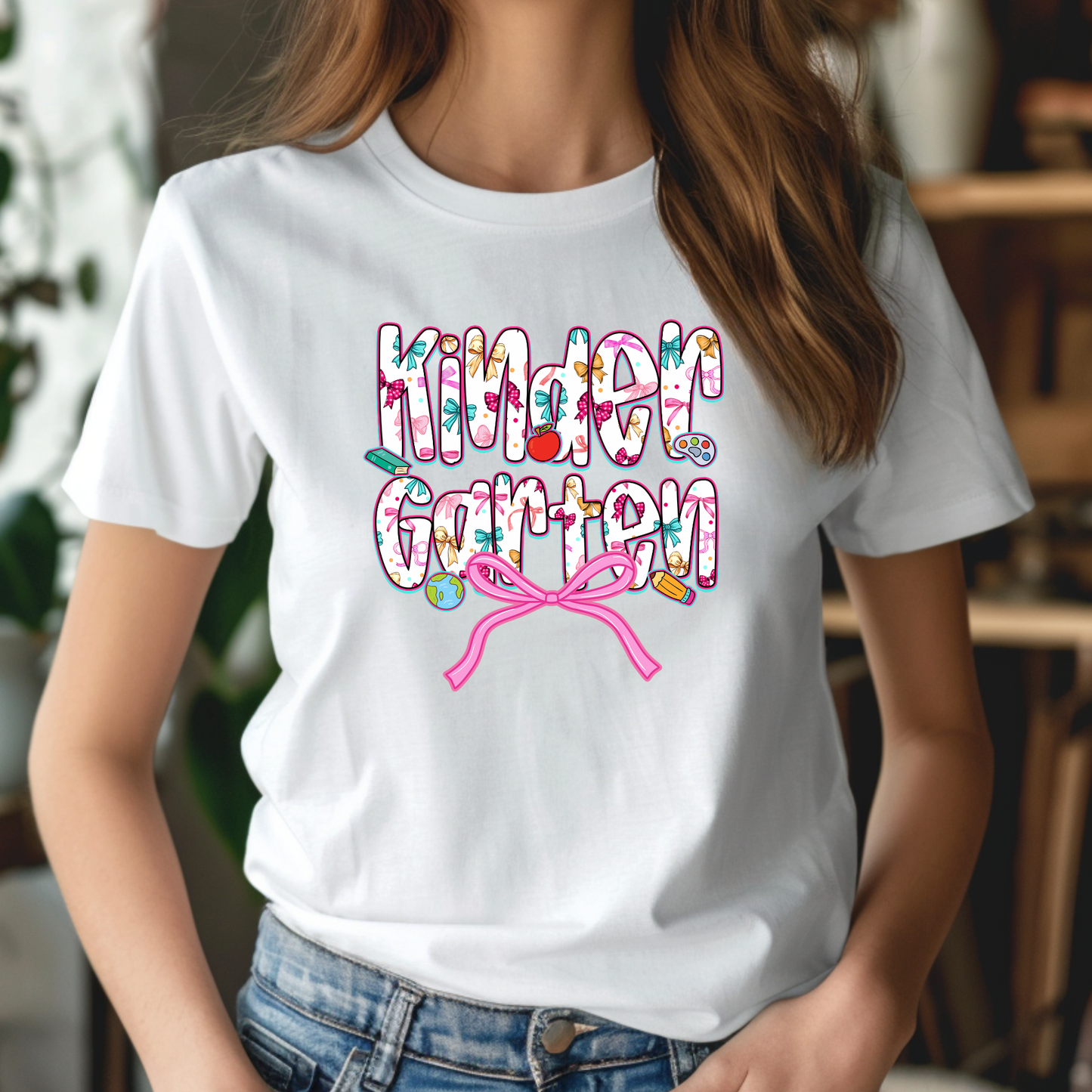 A woman wearing a white short-sleeve graphic T-shirt with the words 'Kinder Garten' in a colorful floral pattern, decorated with small garden motifs and a pink bow, paired with blue jeans.