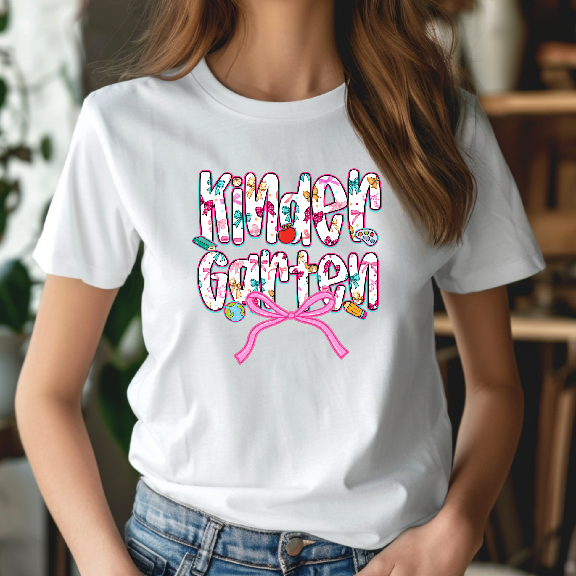 A woman wearing a white short-sleeve graphic T-shirt with the words 'Kinder Garten' in a colorful floral pattern, decorated with small garden motifs and a pink bow, paired with blue jeans.