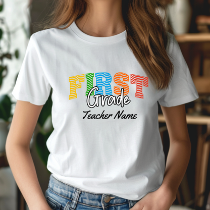 A woman wearing a white short-sleeve graphic T-shirt with the words 'FIRST Grade Teacher Name' printed on the front in colorful and black lettering, paired with blue jeans in a softly blurred indoor setting.