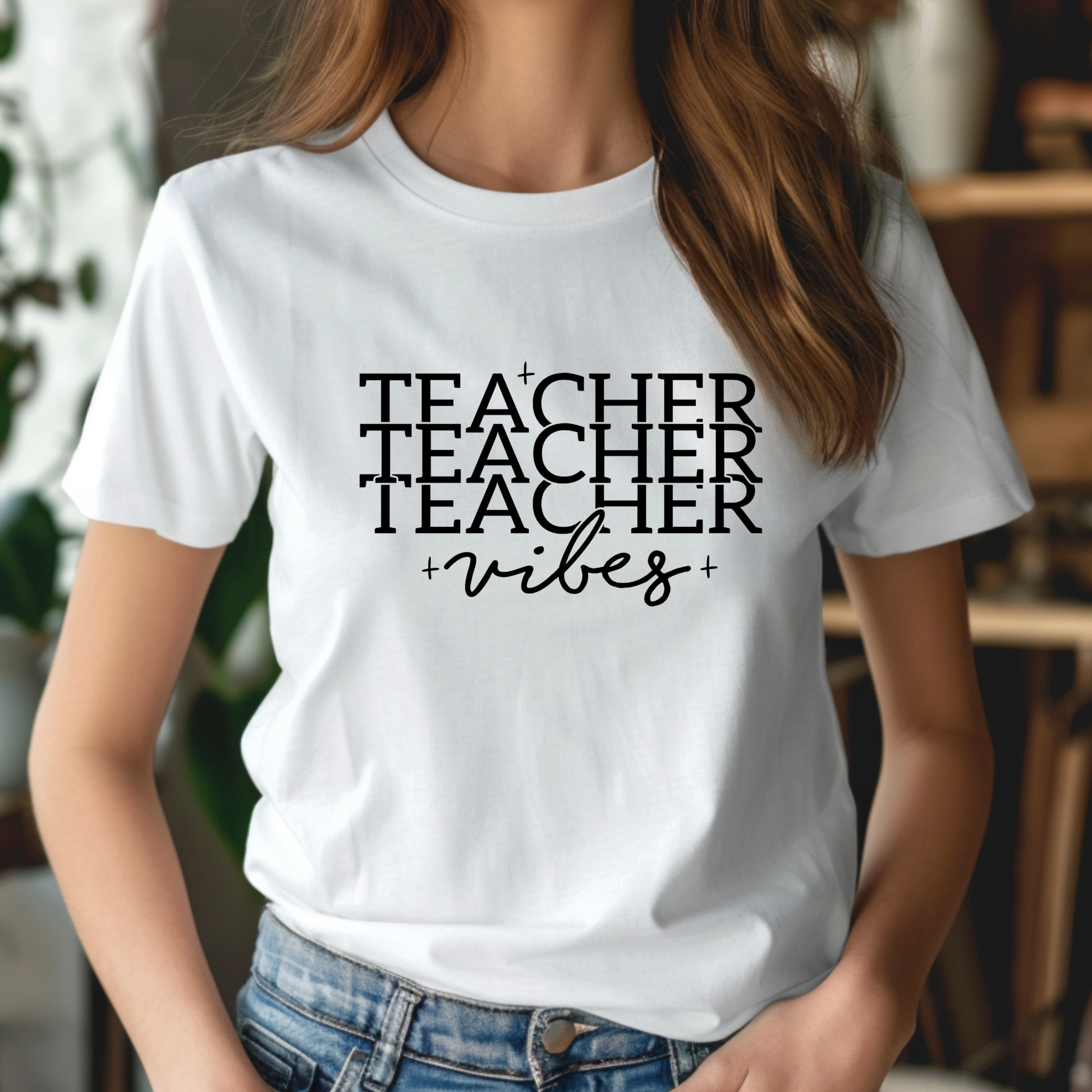 A woman wearing a white short-sleeve crewneck t-shirt with a black graphic text design that reads 'TEACHER TEACHER TEACHER +vibes+' in a stacked, layered font. The photo is cropped from the neck down, showing her brown hair, the t-shirt, and blue jeans against a softly blurred indoor background.
