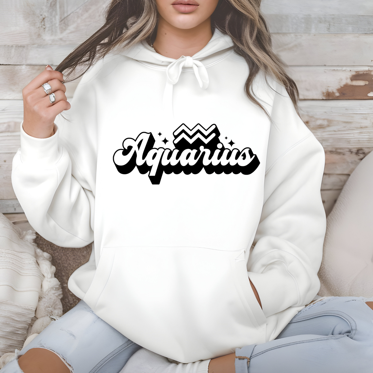 A woman wearing a white pullover hoodie with a black 'Aquarius' graphic on the chest, sitting against a rustic wood and beige cushion background.