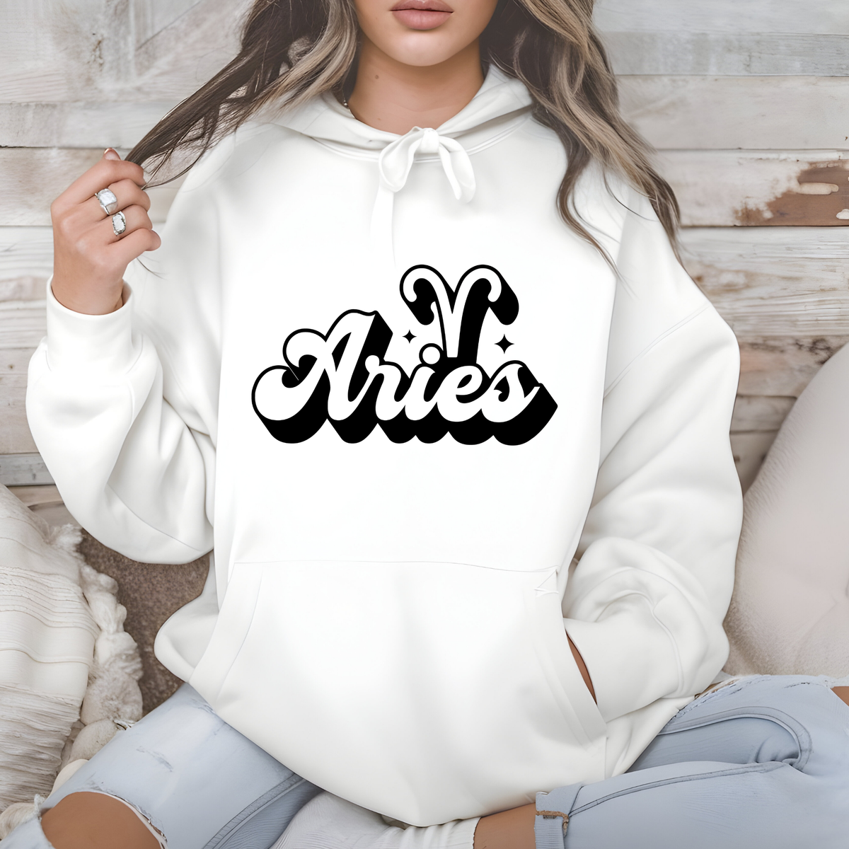 A woman wearing a white pullover hoodie with a black 'Aries' zodiac sign graphic on the chest, sitting against a rustic wood panel background.