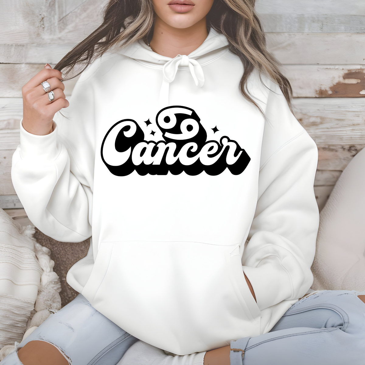 A woman wearing a white pullover hoodie with a black 'Yo Capricorn' graphic on the chest, sitting against a rustic wood panel background.